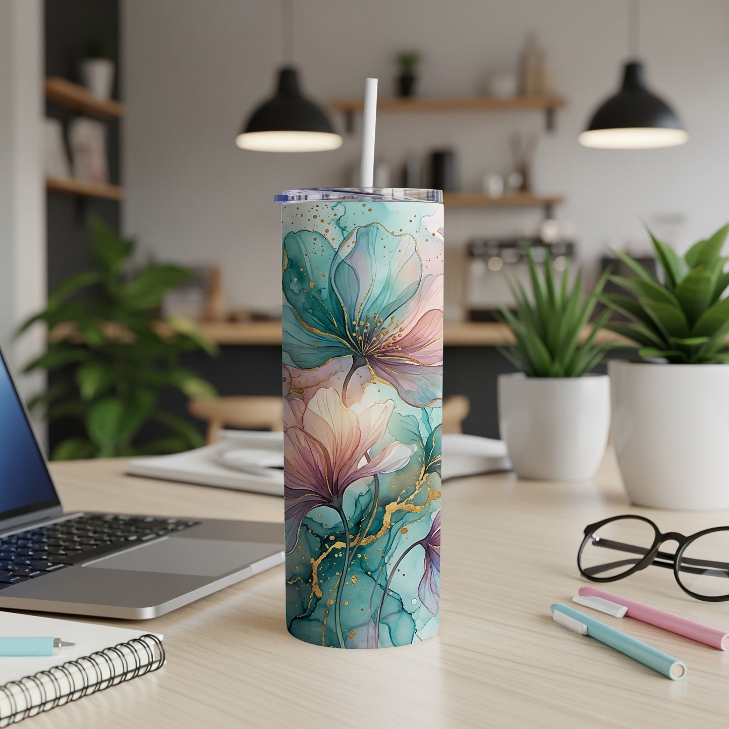 Floral tumbler with straw on a desk next to a laptop and pens.
