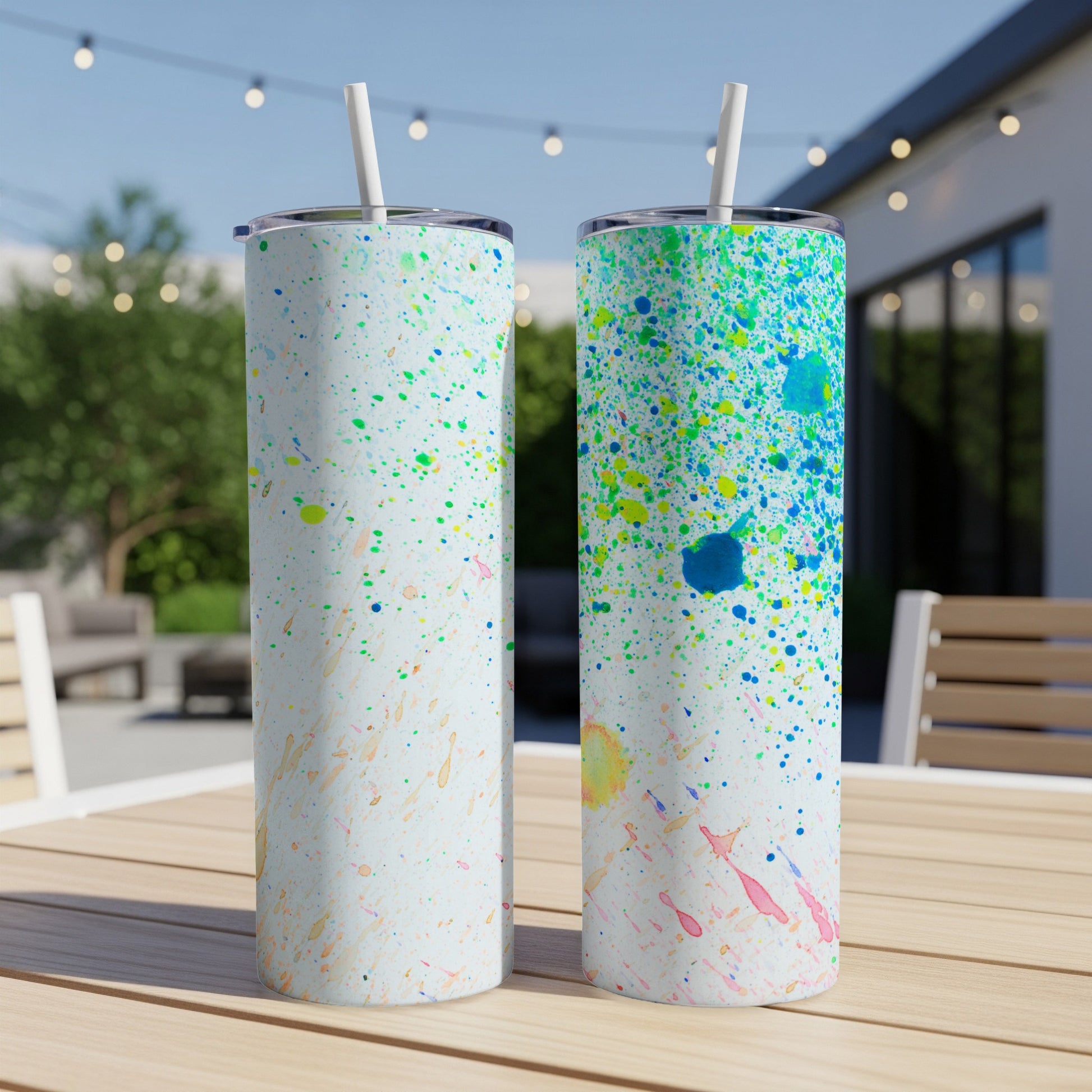 Two tumblers with colorful paint splatters on a wooden table outdoors.