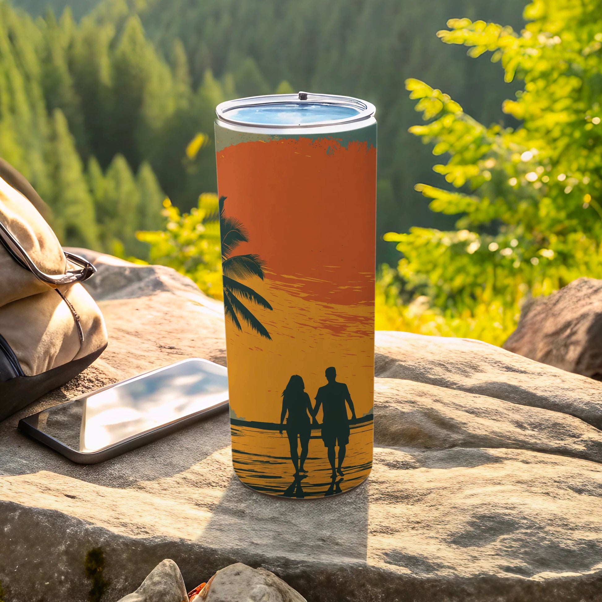 Tumbler with sunset beach silhouette of couple walking, phone, and bag on rocks.
