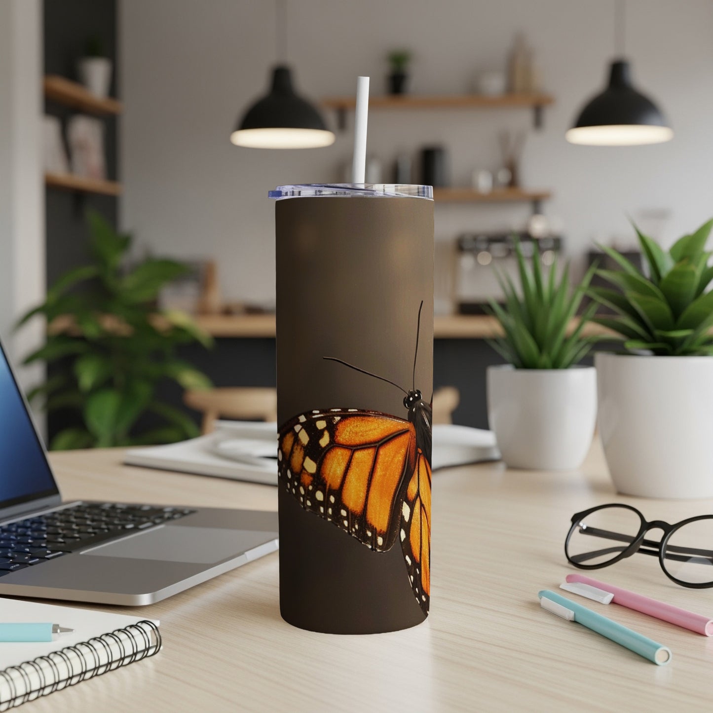 Monarch butterfly on a brown tumbler with a straw, laptop, and pens.