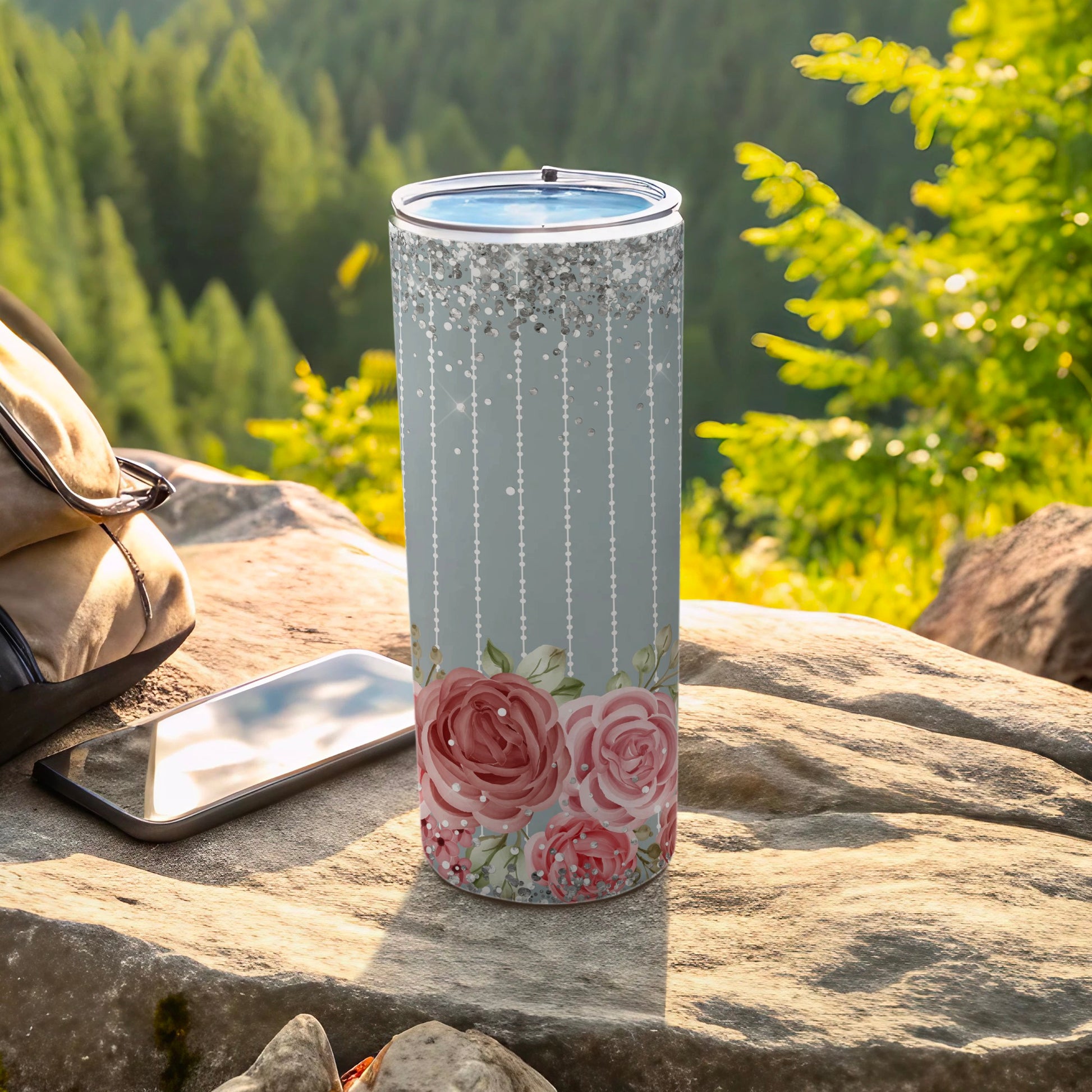 Gray tumbler with pink roses, glitter border, on rocky terrain with backpack.
