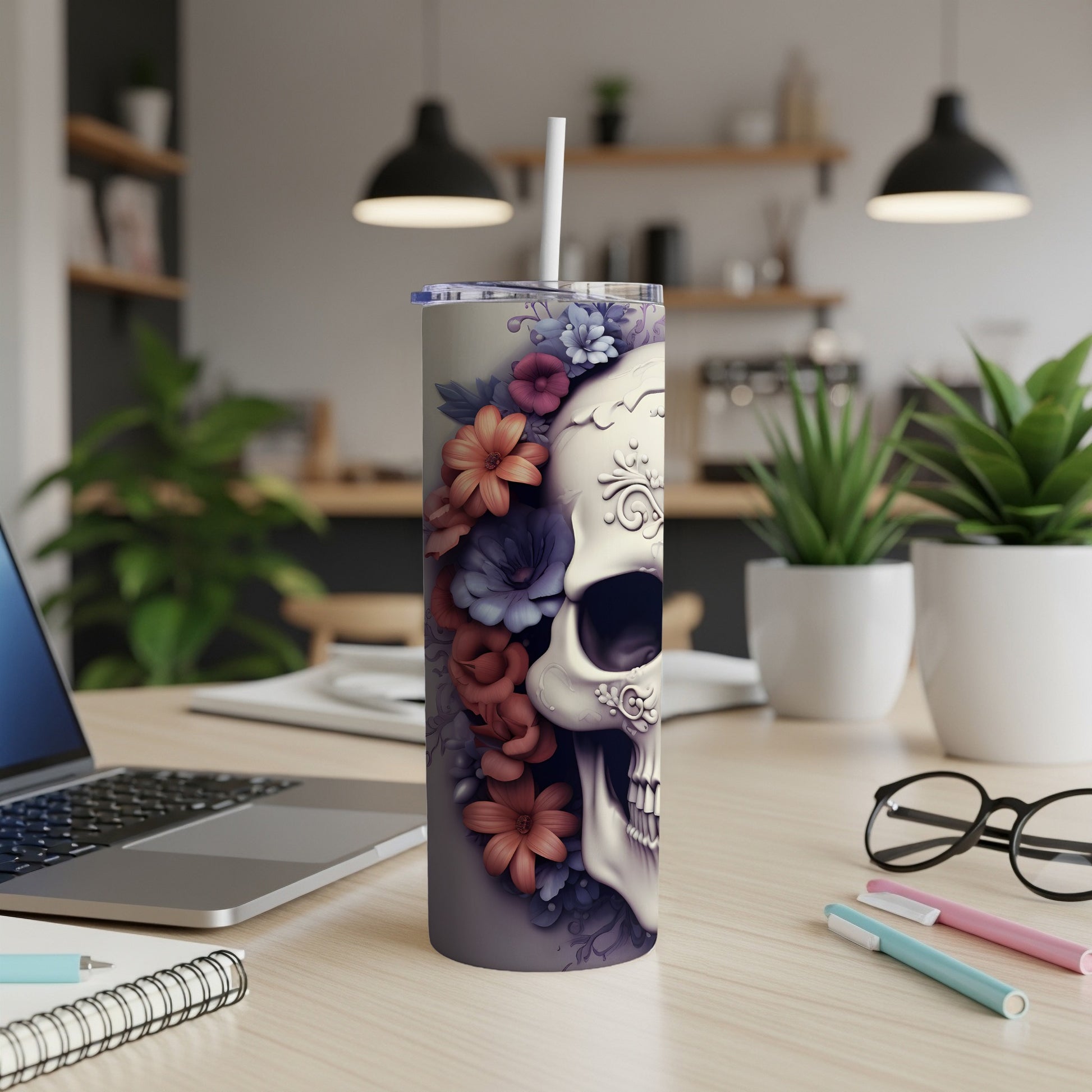 Floral skull tumbler with straw on a desk with laptop and pens.