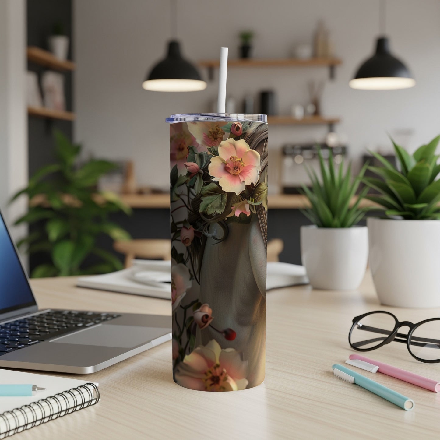Floral tumbler with a straw on a wooden desk next to a laptop and pens.