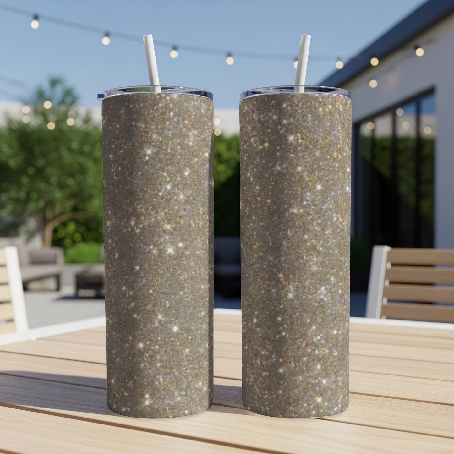 Two glitter-covered tumblers with straws sit on a wooden table outdoors.