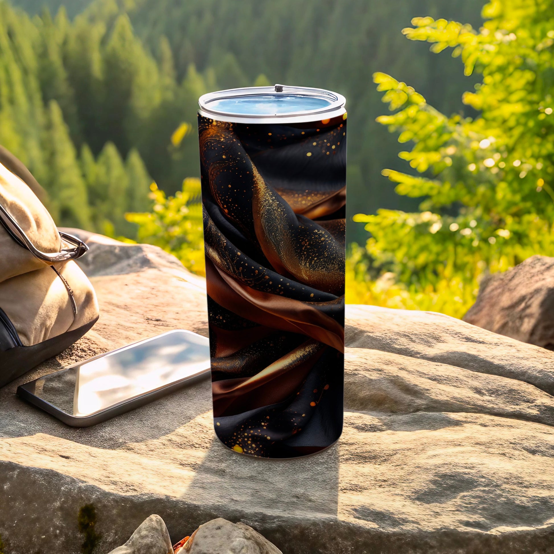 Tumbler with dark fabric and gold glitter design on rock with backpack and phone.