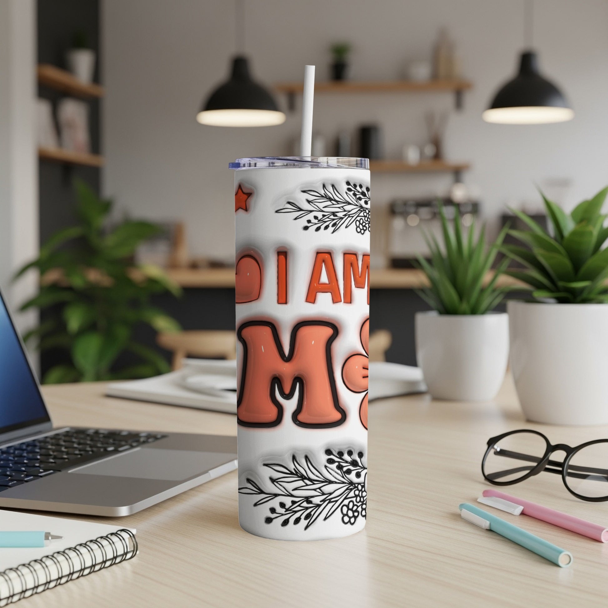 White tumbler with "I AM MOM" text and floral accents on a desk.