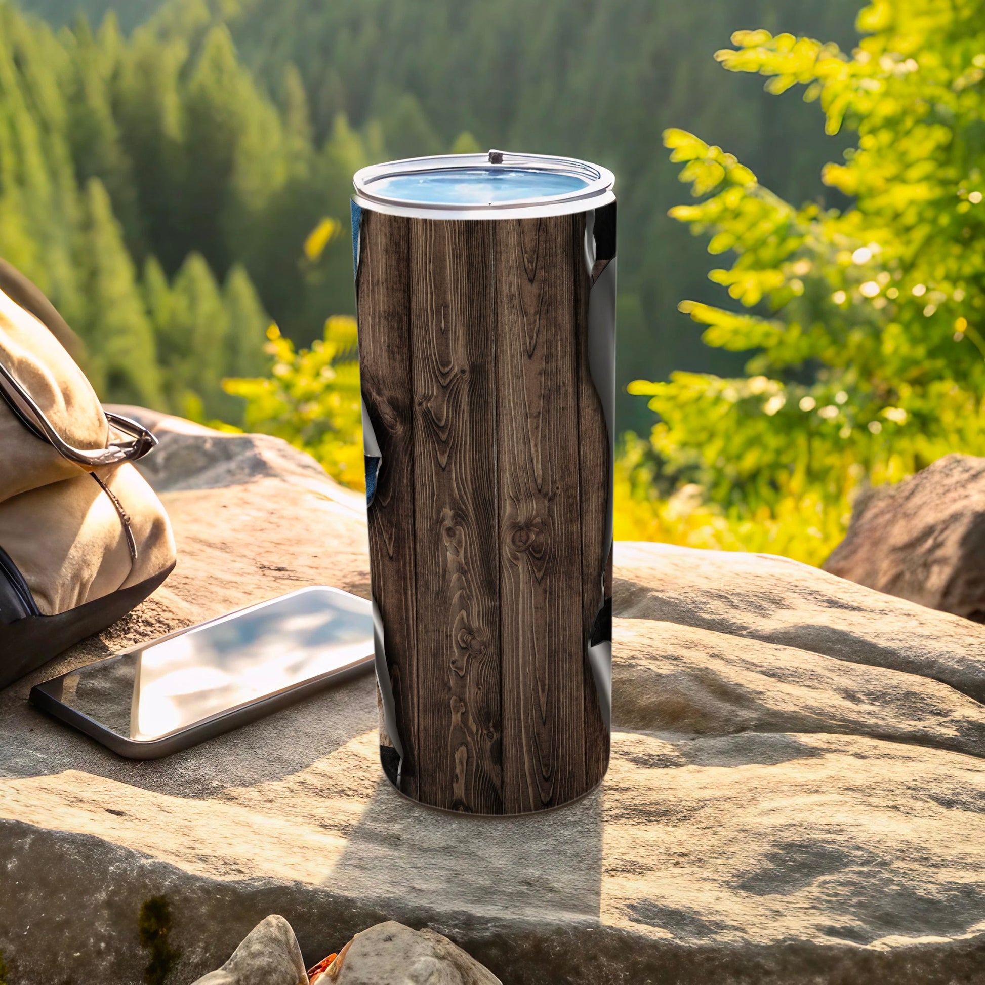 Tall tumbler with wood grain design, on rock with phone and bag.