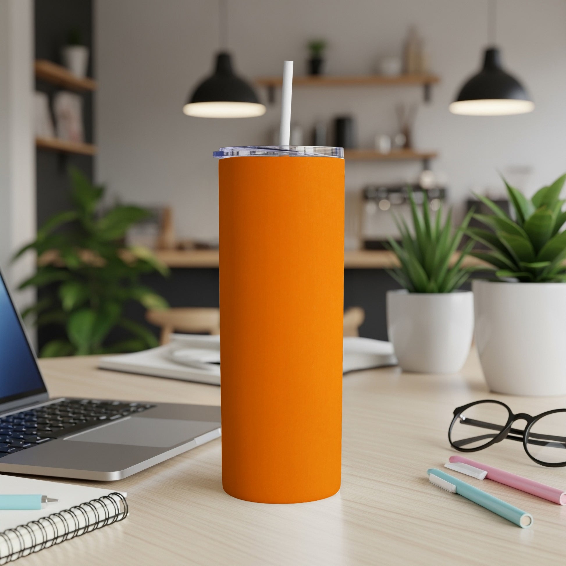 Orange tumbler with straw on a desk next to a laptop and pens.