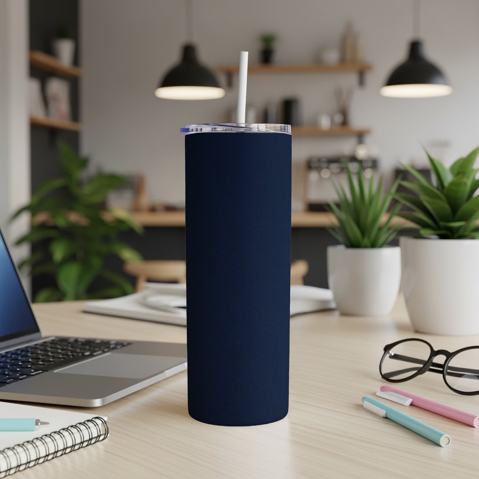 Dark blue tumbler with white straw on a desk with laptop and pens.