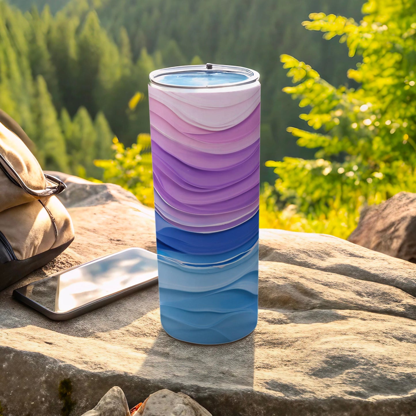 Tumbler with wave pattern of purple and blue layers on rock with phone and backpack.