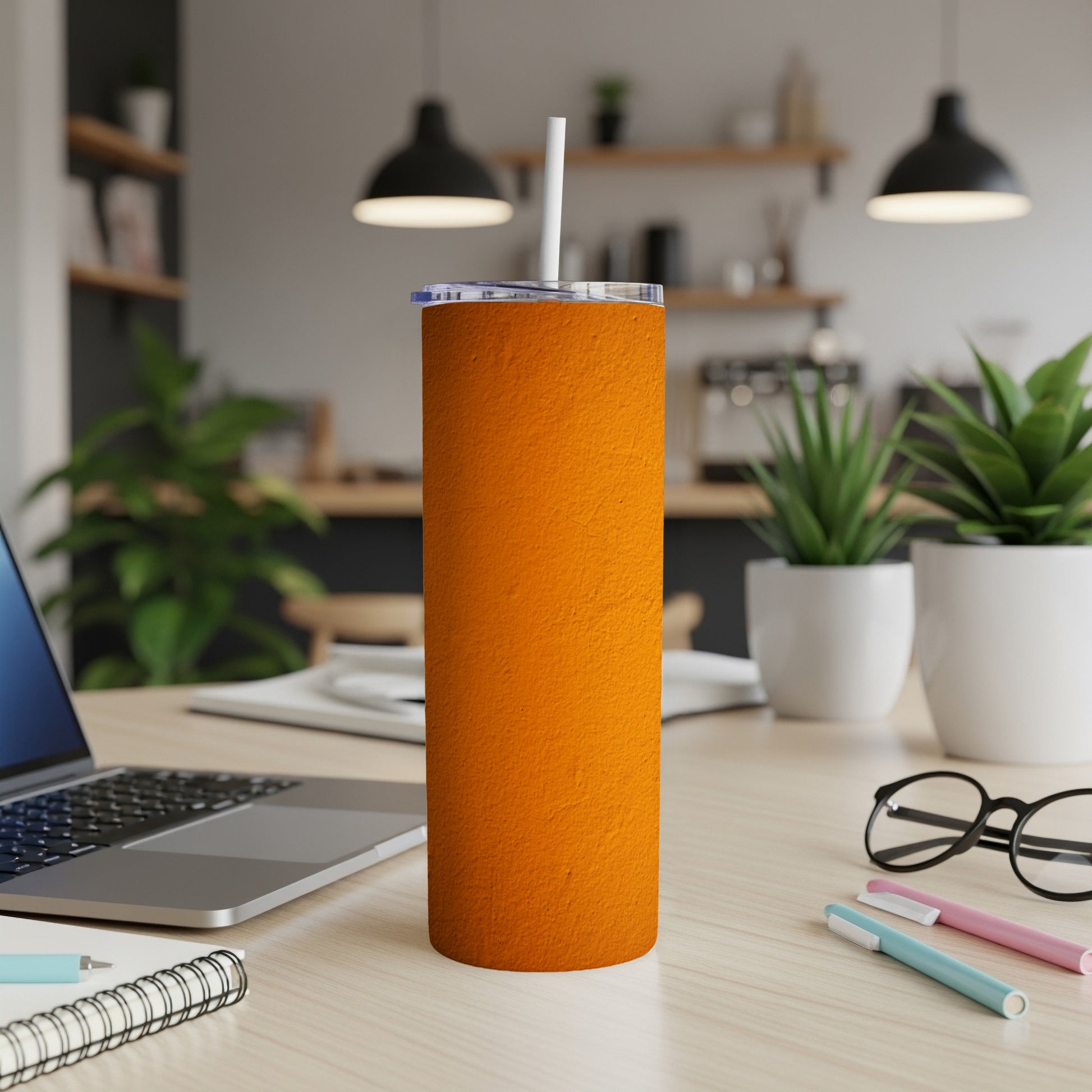 Orange textured tumbler with white straw on a desk with laptop and pens.