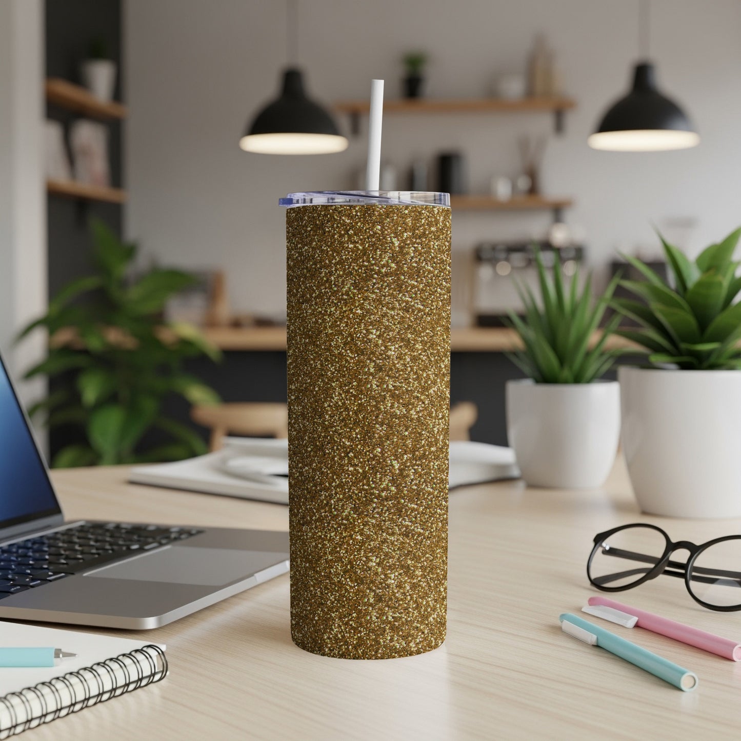 Gold glitter tumbler with straw on a desk beside a laptop and notepad.