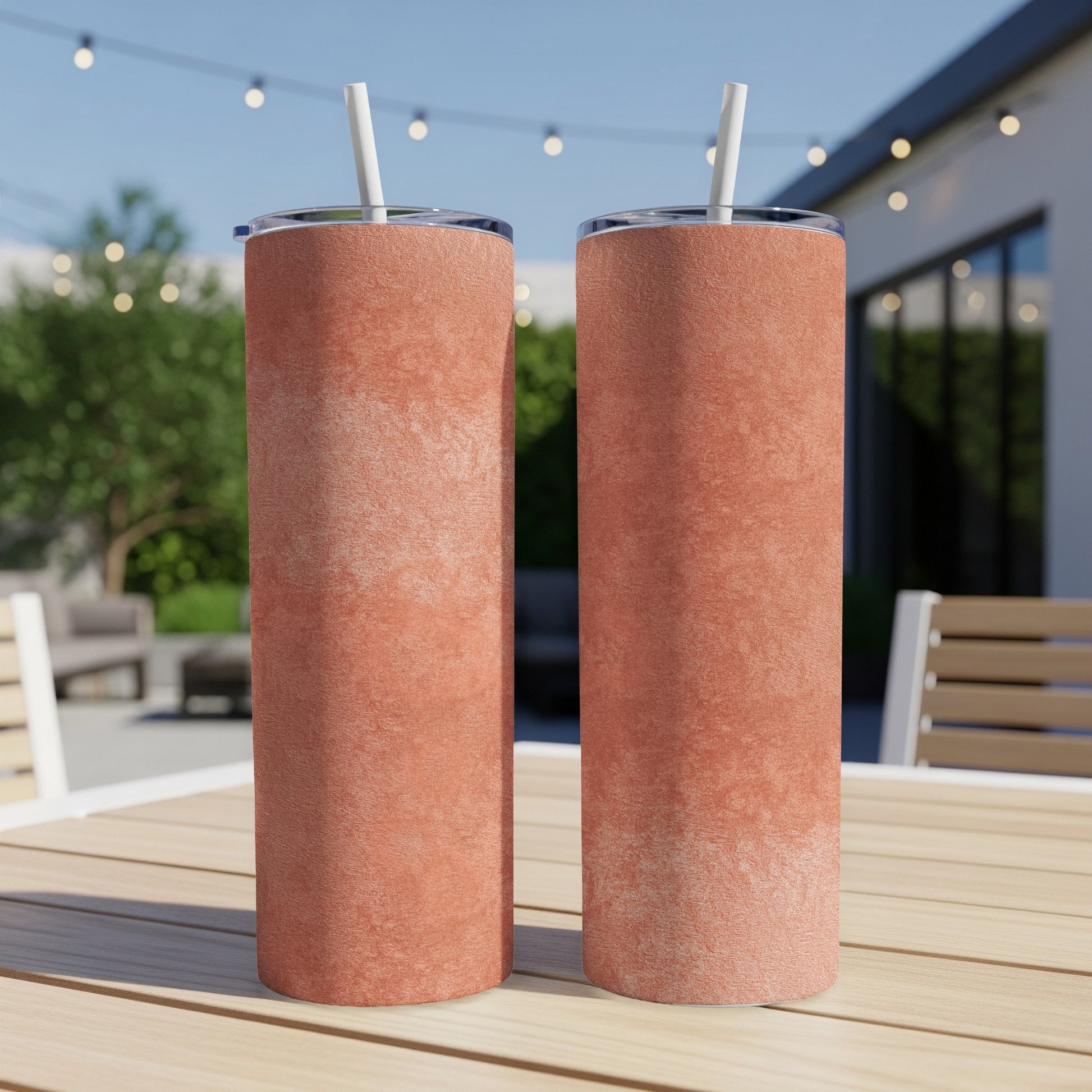 Two salmon-colored tumblers with white straws stand on a wooden table outdoors.