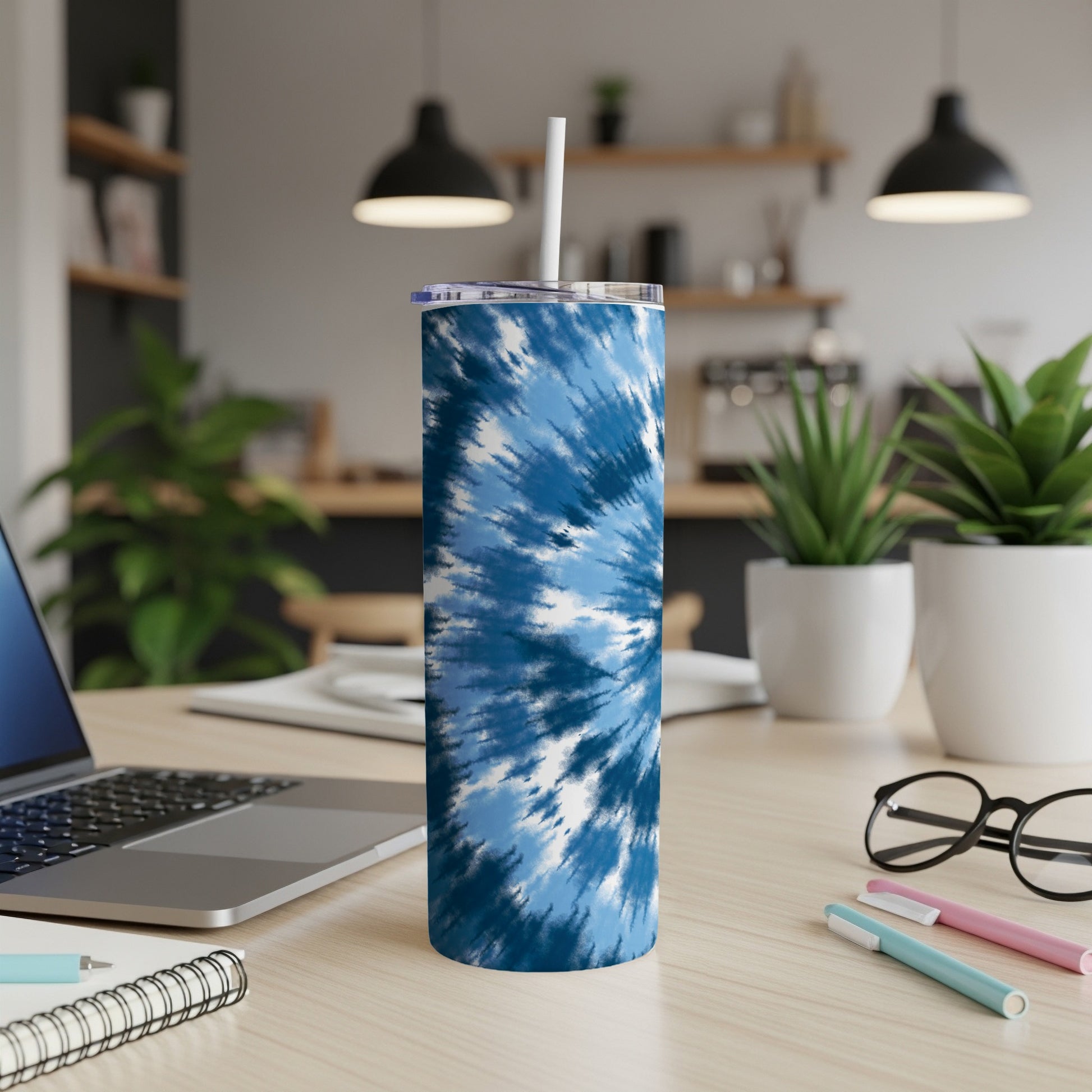 Blue tie-dye tumbler with straw on a wooden desk next to a laptop.