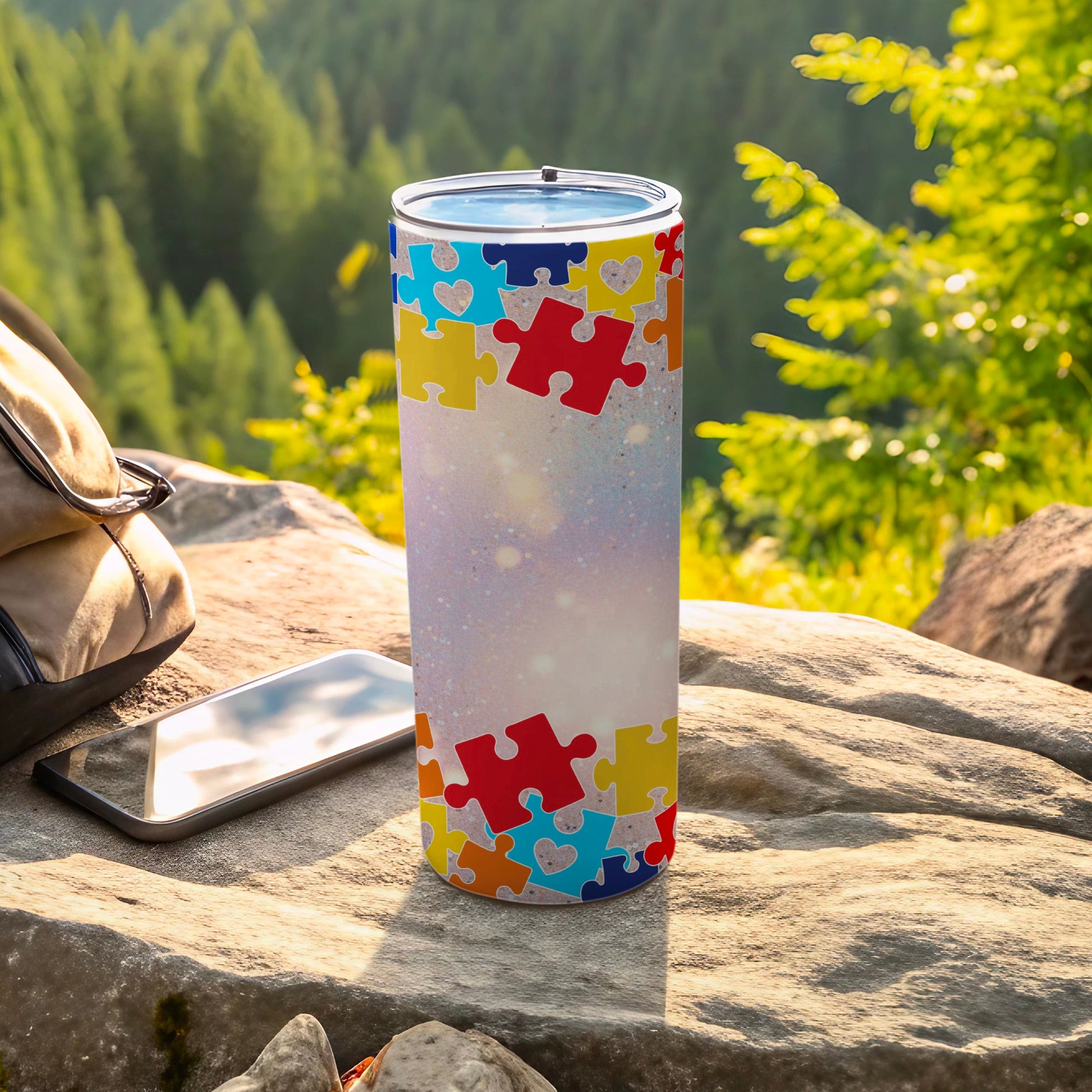 Tumbler with colorful puzzle pieces pattern, resting on rocks with phone and backpack.