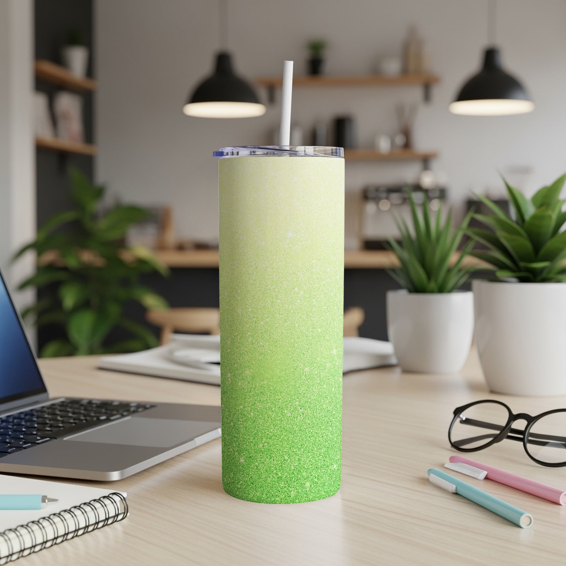 Green glitter tumbler with straw on a wooden desk with laptop and pens.