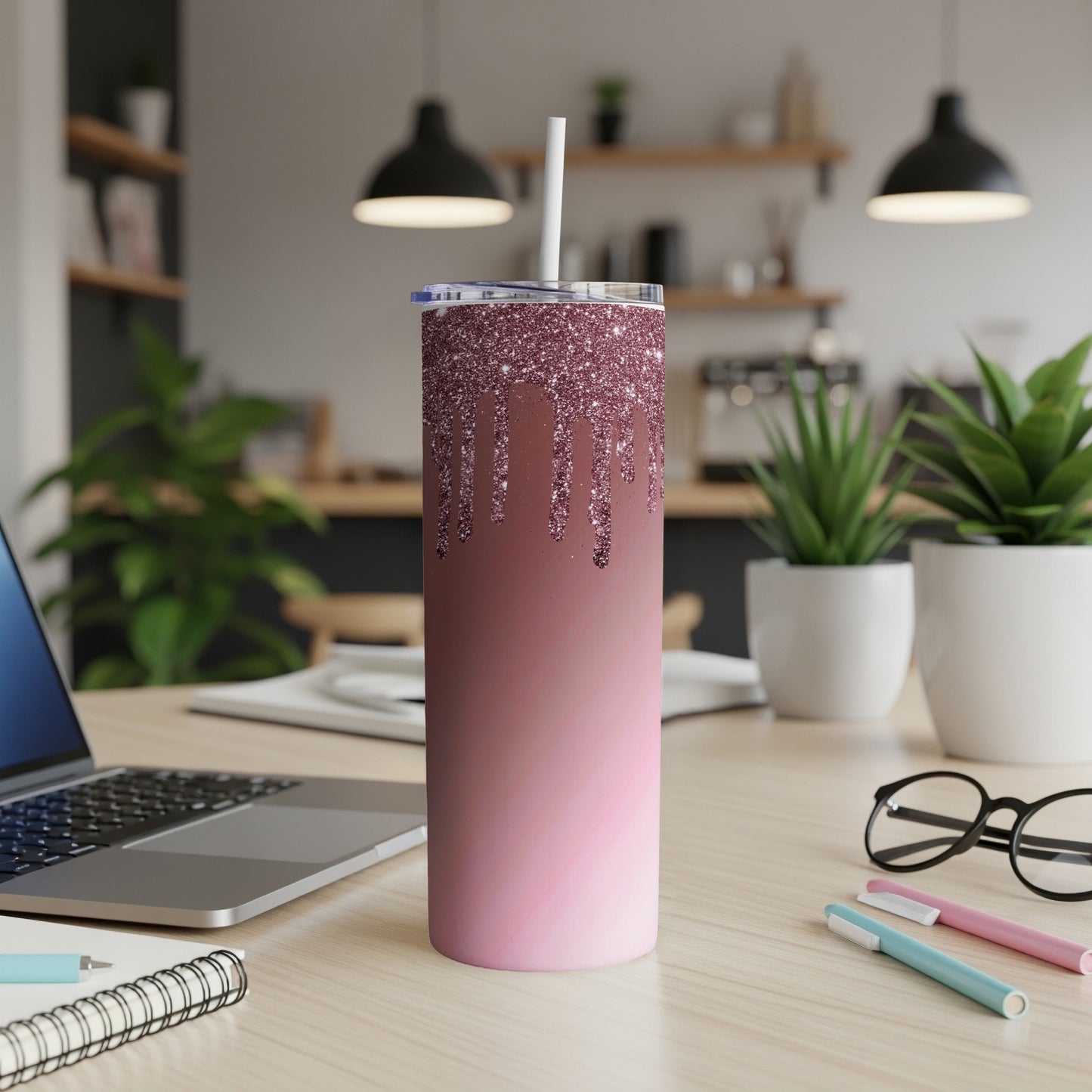 Glitter drip-effect skinny tumbler with straw on a wooden desk with laptop and plants.