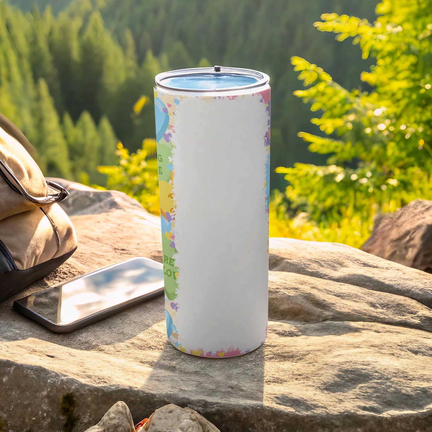 White tumbler with colorful confetti pattern sits on rock with phone and backpack.