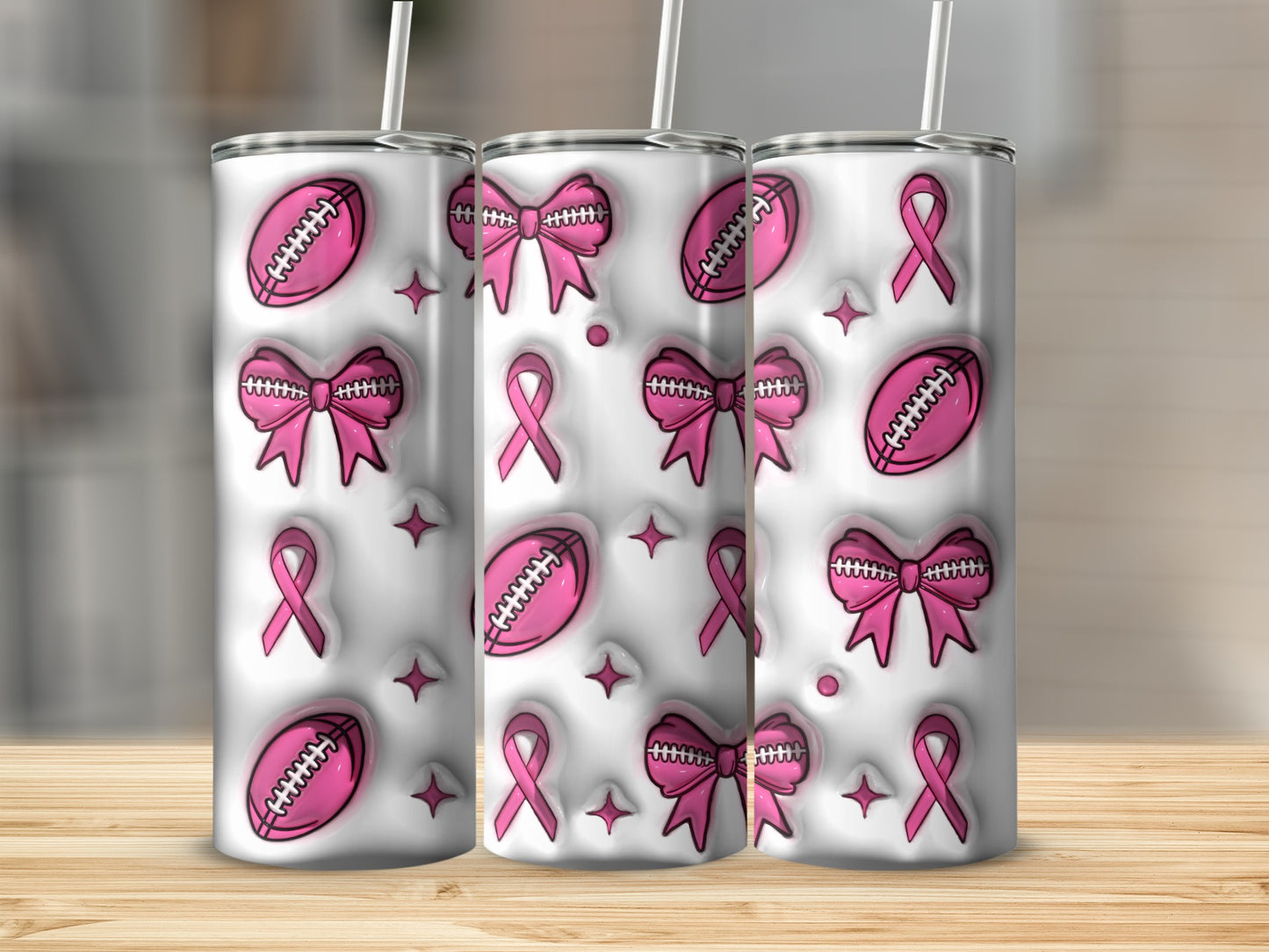 Three pink ribbon and football themed tumblers with straws on a wooden surface.