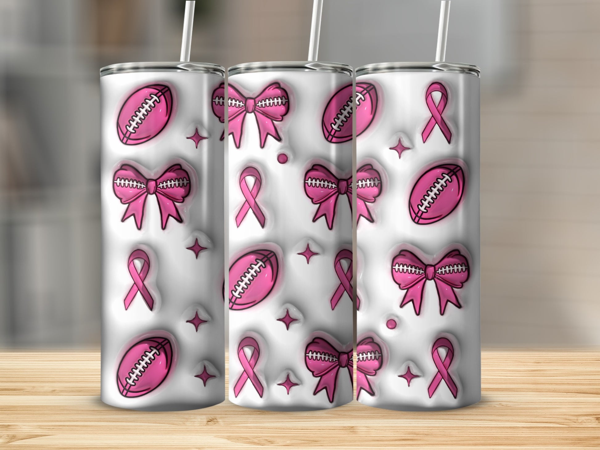Three pink ribbon and football themed tumblers with straws on a wooden surface.