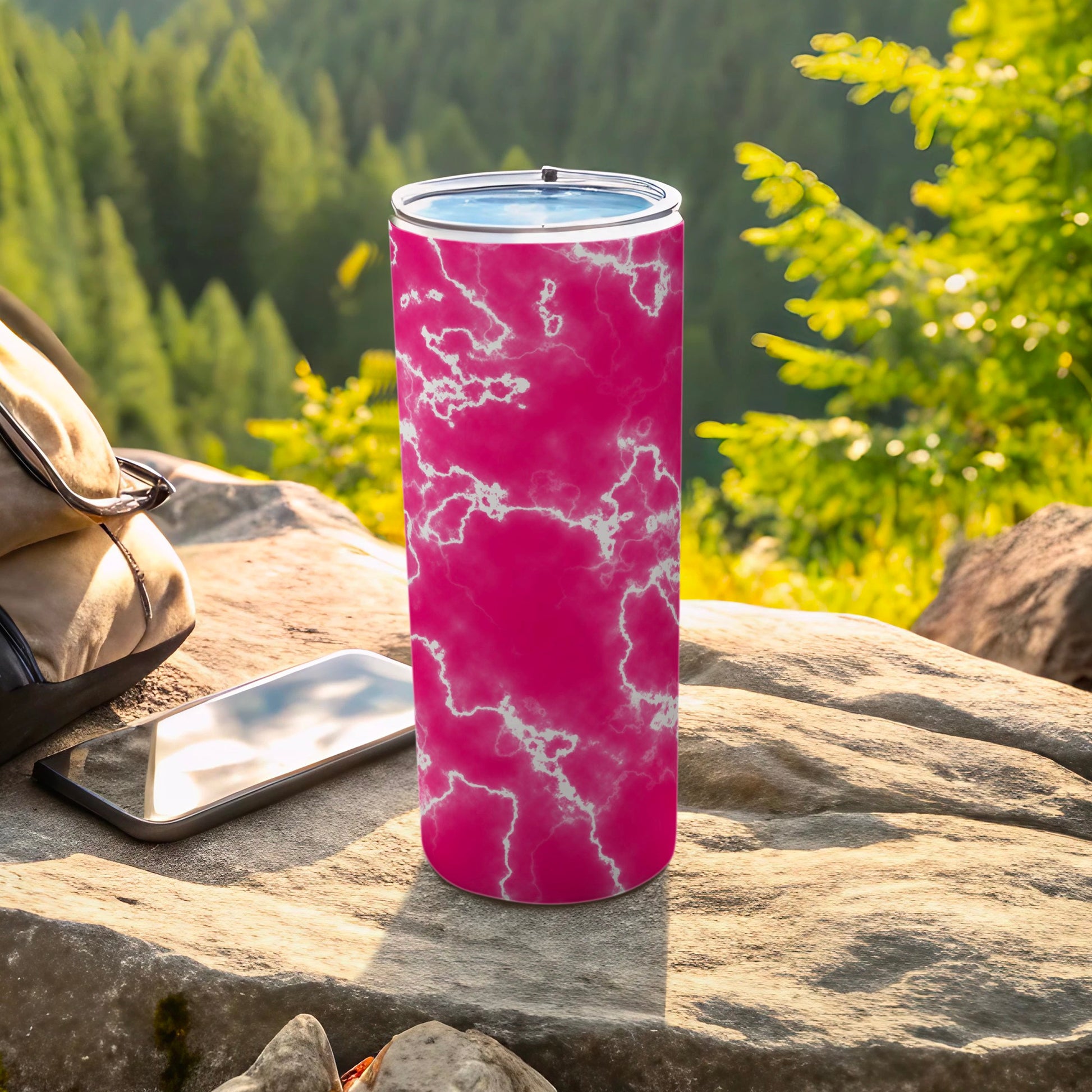 Pink marble pattern tumbler with lid on rocky surface with phone and backpack.