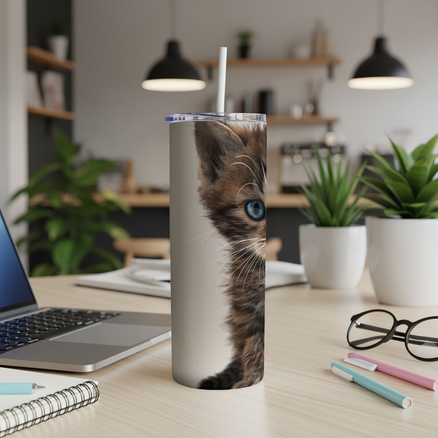 Tabby kitten with blue eyes peeking from a tumbler on a desk.