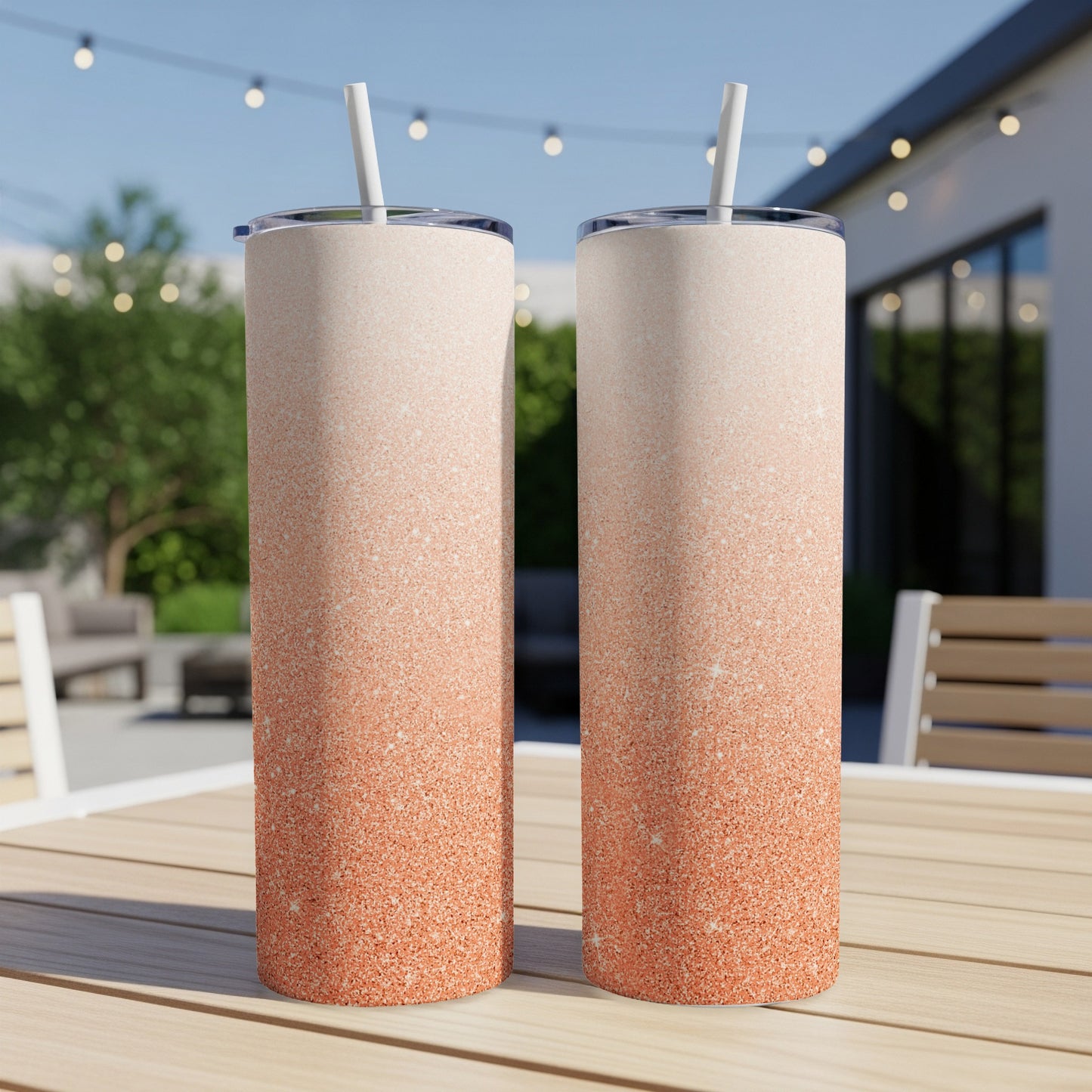 Two rose gold glitter gradient tumblers with straws on a wooden table outdoors.