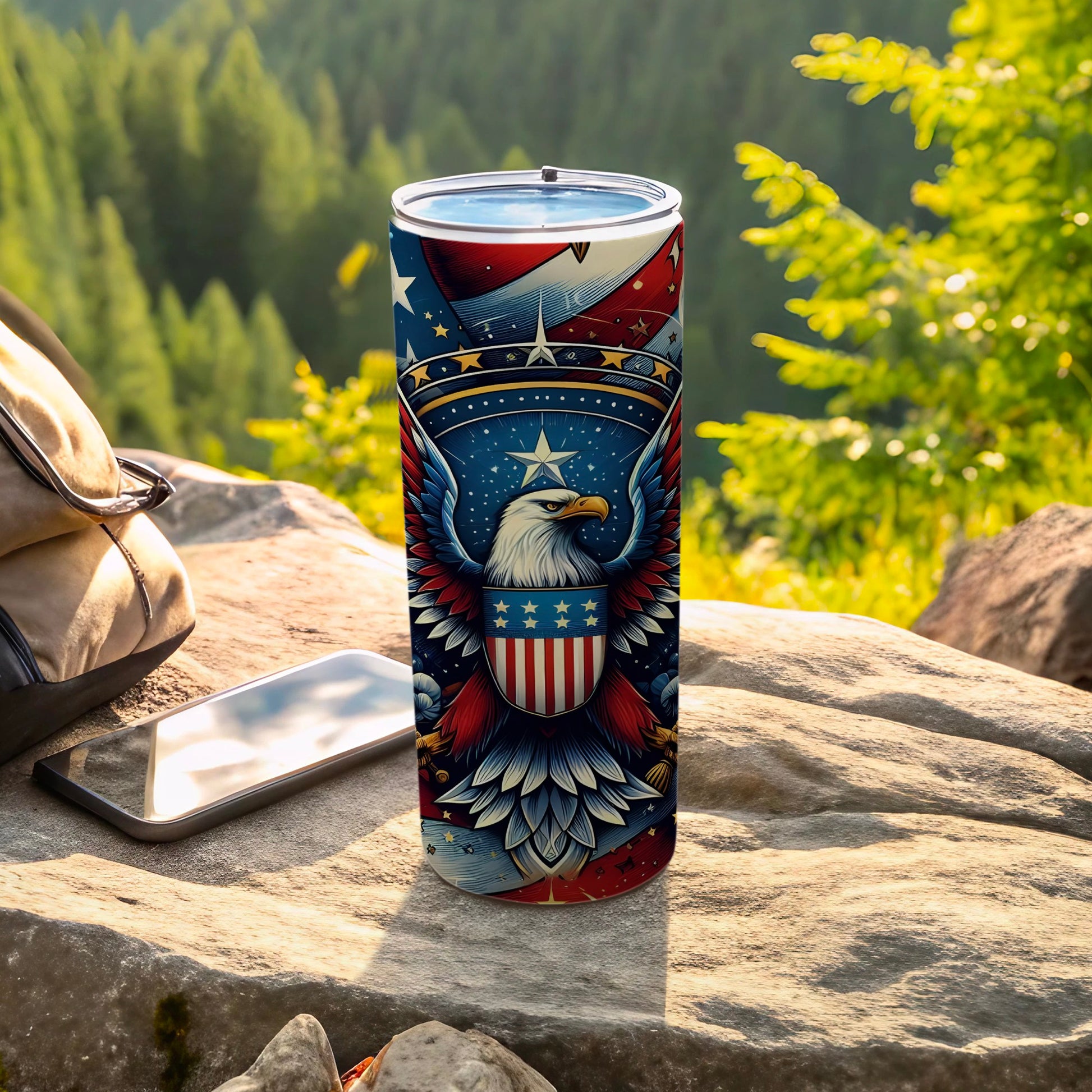 Patriotic tumbler with eagle and stars, set on rock with backpack and phone.