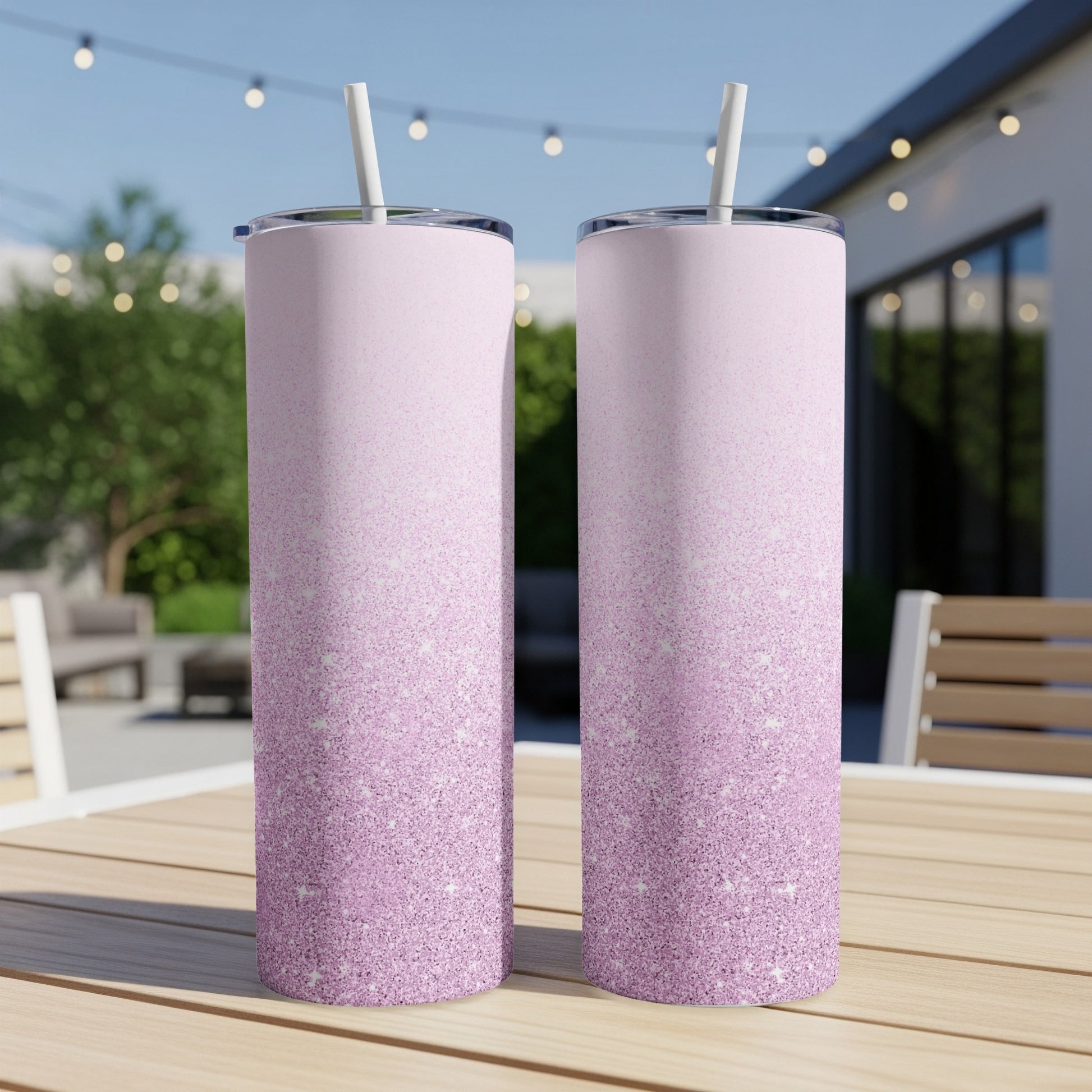 Two glittery pink ombre tumblers with straws on a wooden table outdoors.