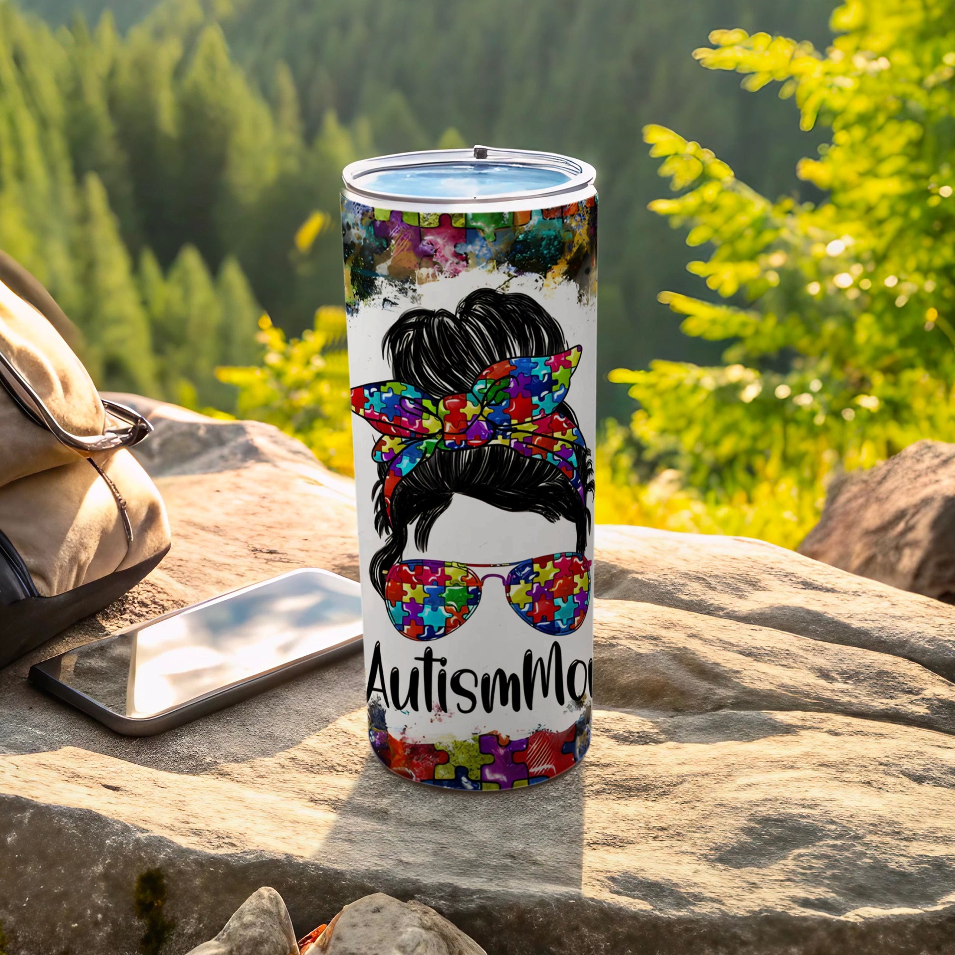Tumbler with Autism mom design, puzzle pieces, messy bun, sunglasses, on a rock.