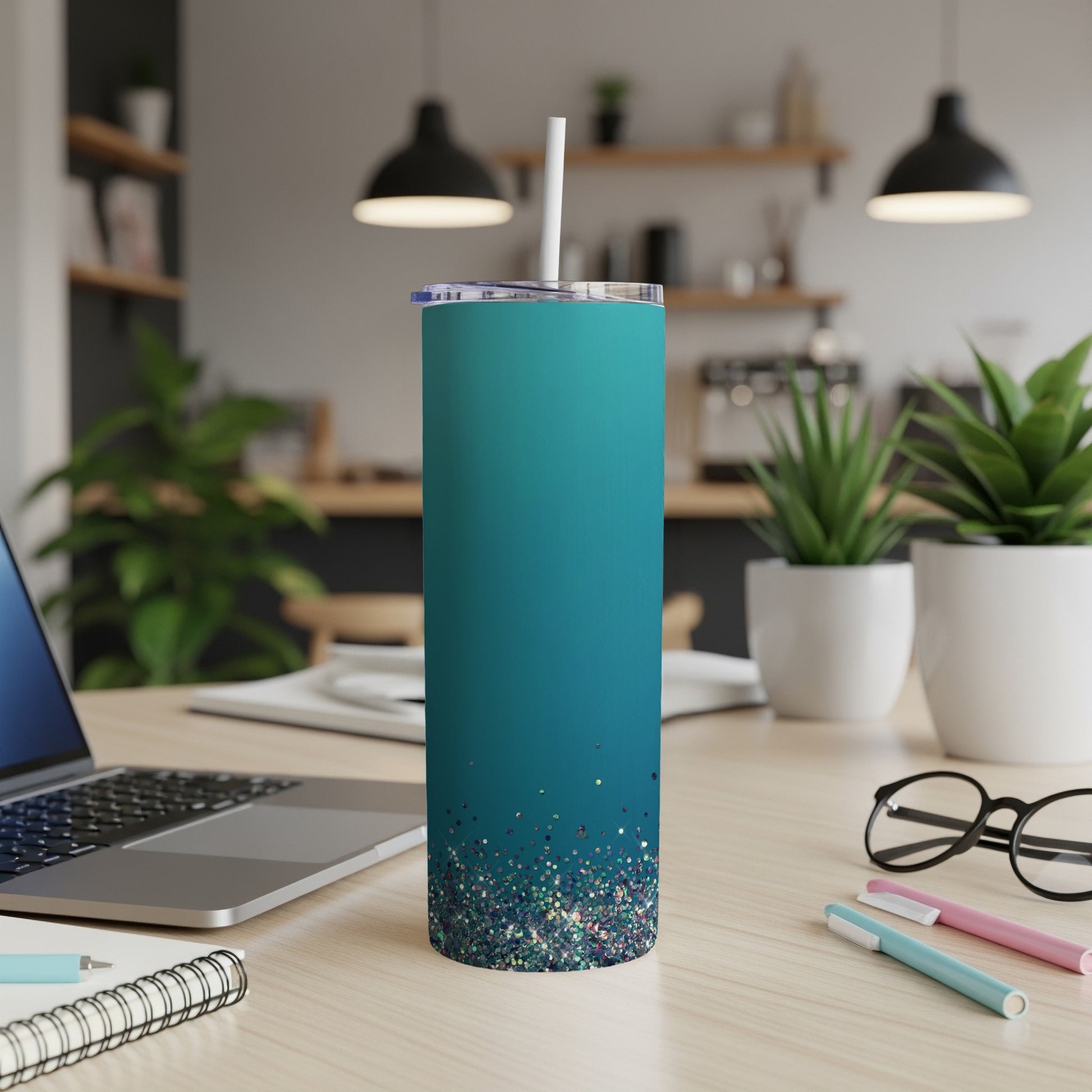 Teal tumbler with glitter bottom and straw on a wooden desk.