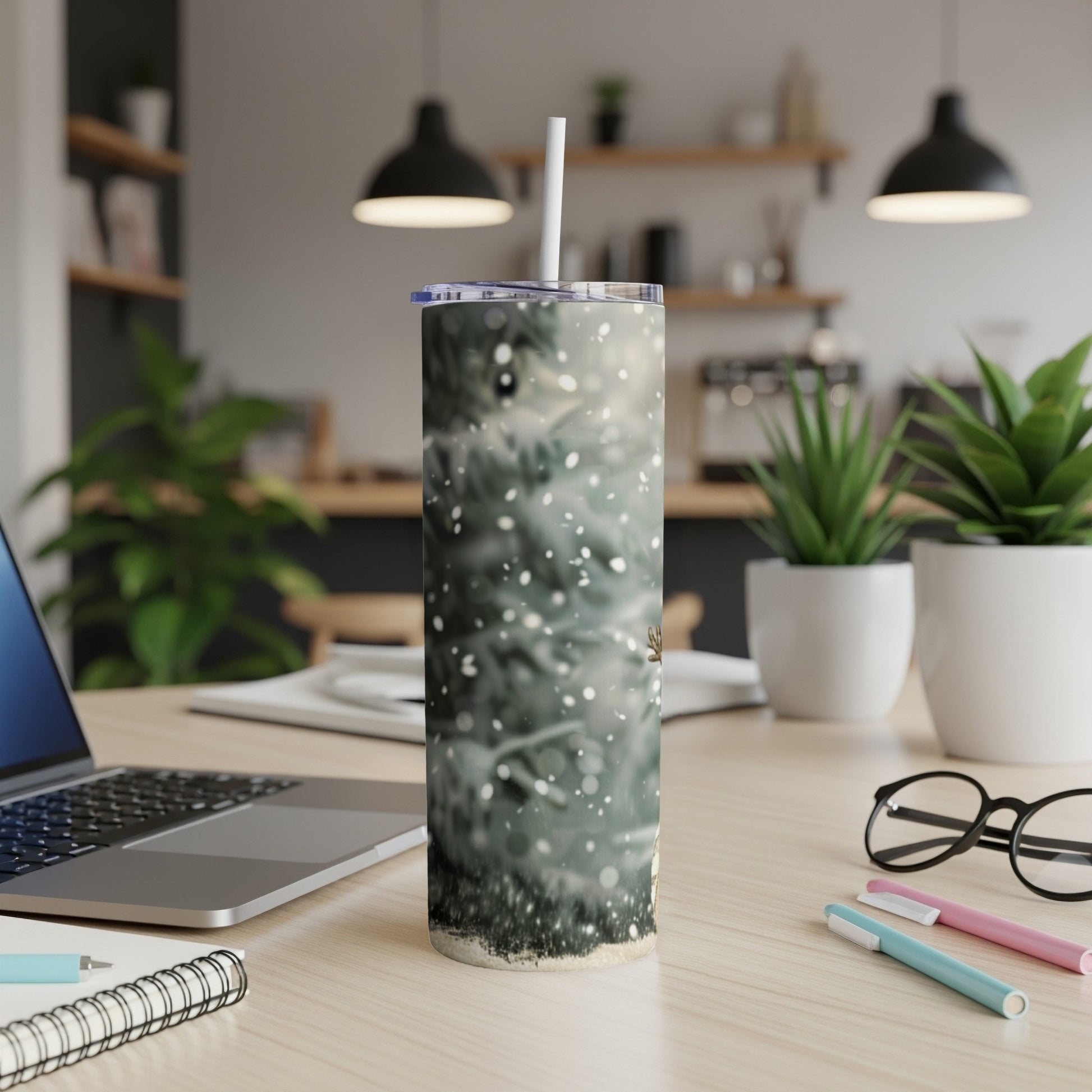Tumbler with snowy evergreen trees and subtle bokeh, on a desk with a laptop.