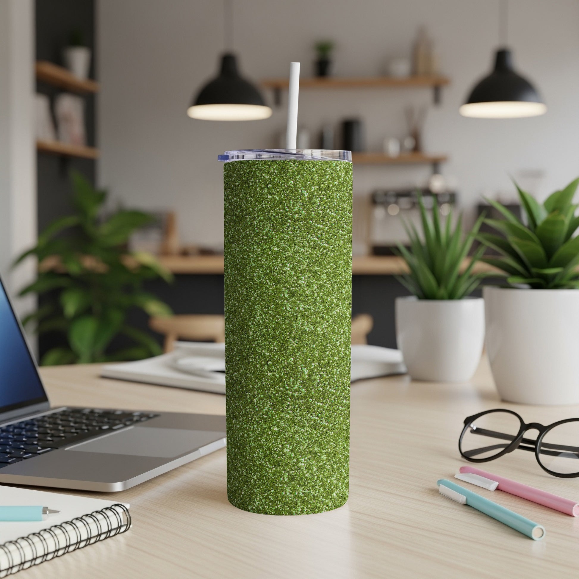 Green glitter tumbler with straw on a wooden desk with laptop and supplies.