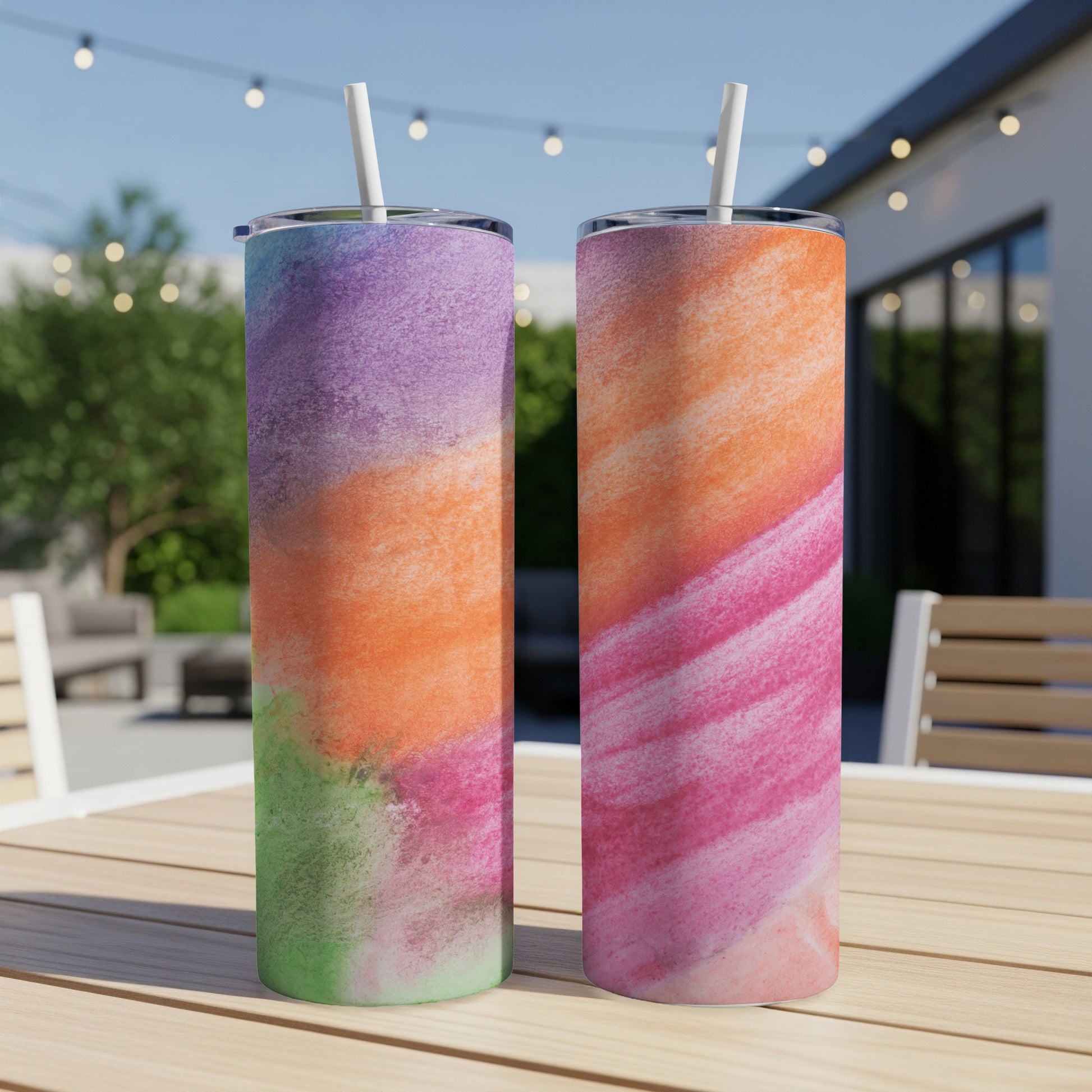 Two tumblers with abstract watercolor designs sit on a wooden table outdoors.