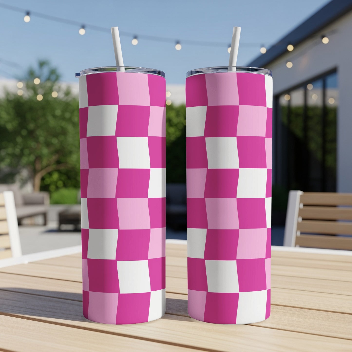 Two pink and white checkered tumblers with straws sit on a wooden table outdoors.