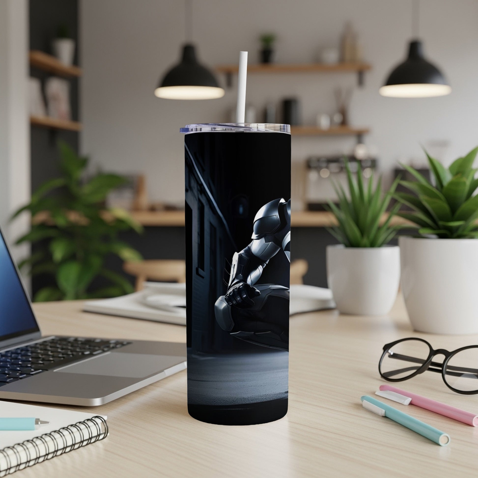 Dark gray tumbler with robot graphic on desk with laptop and pens.