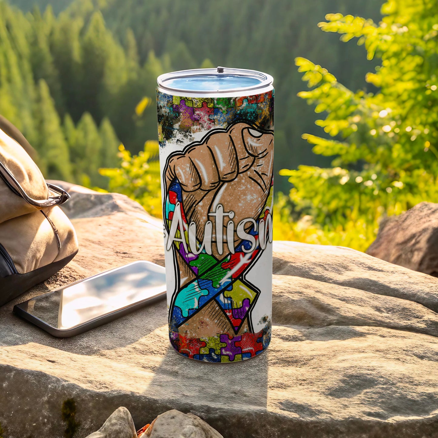 Tumbler with autism puzzle ribbon fist design, resting on rocks.