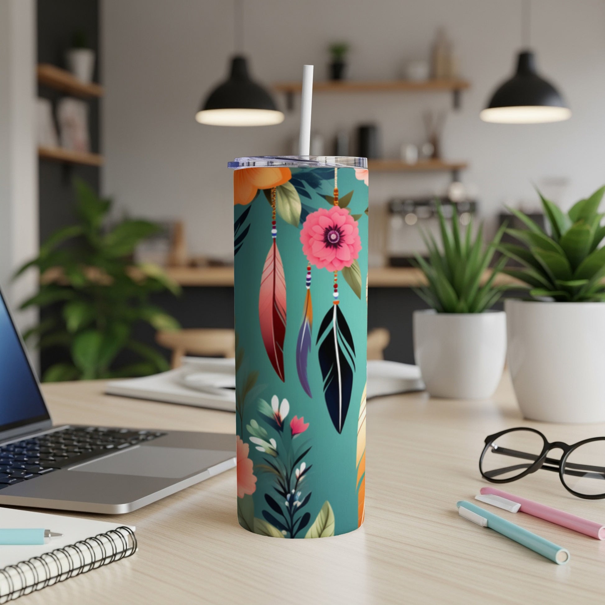 Teal tumbler with floral and feather design, straw, on desk with laptop and pens.
