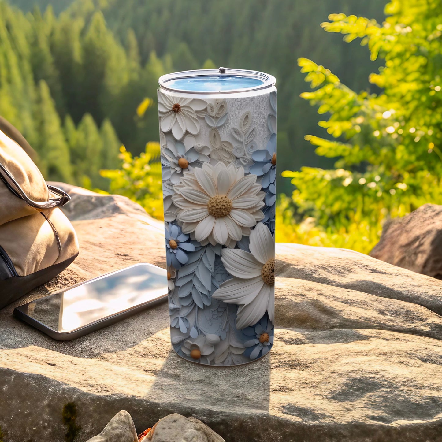Tall white and blue floral embossed tumbler on rock with phone and backpack.