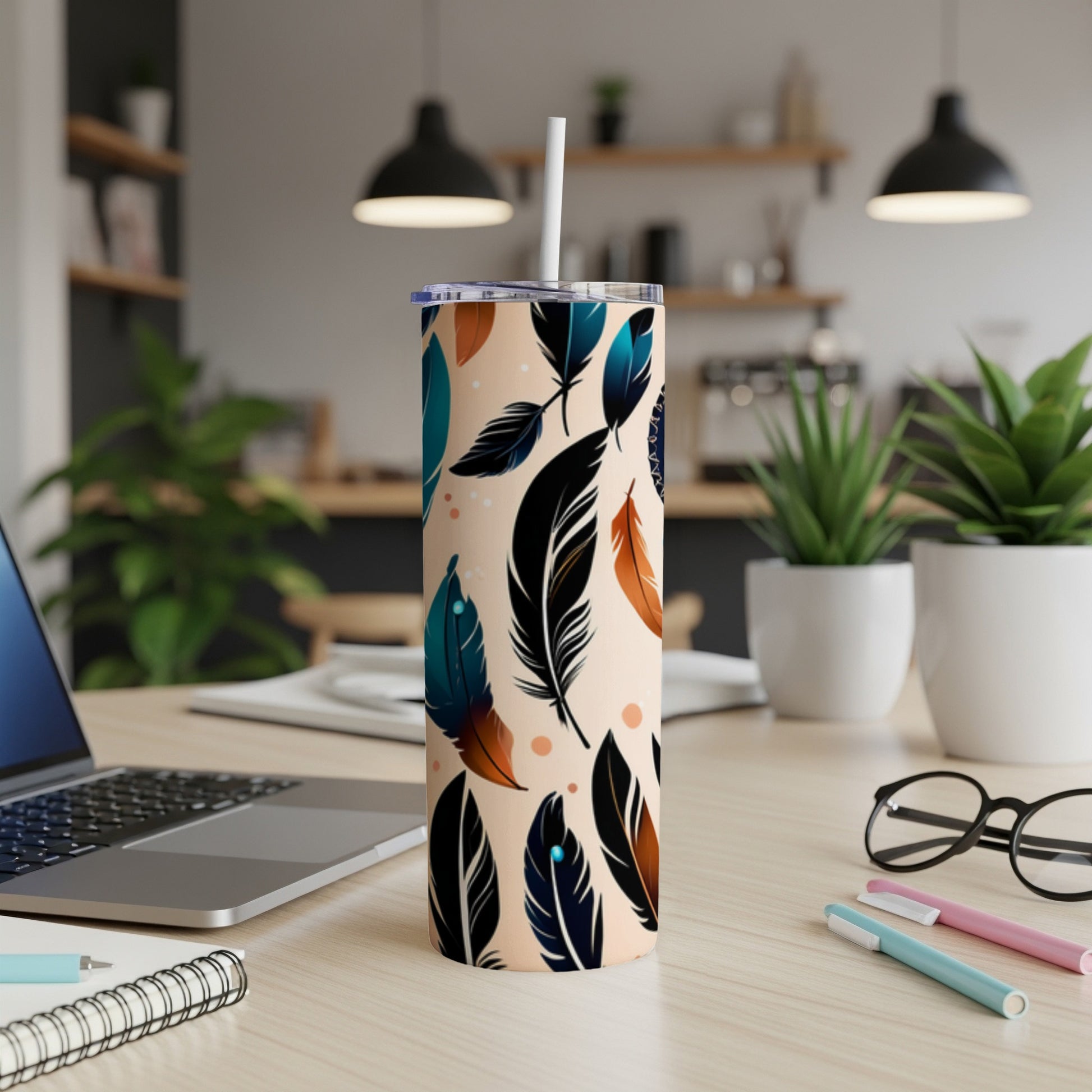 Tall tumbler with repeating feather pattern, straw, laptop, and pens.