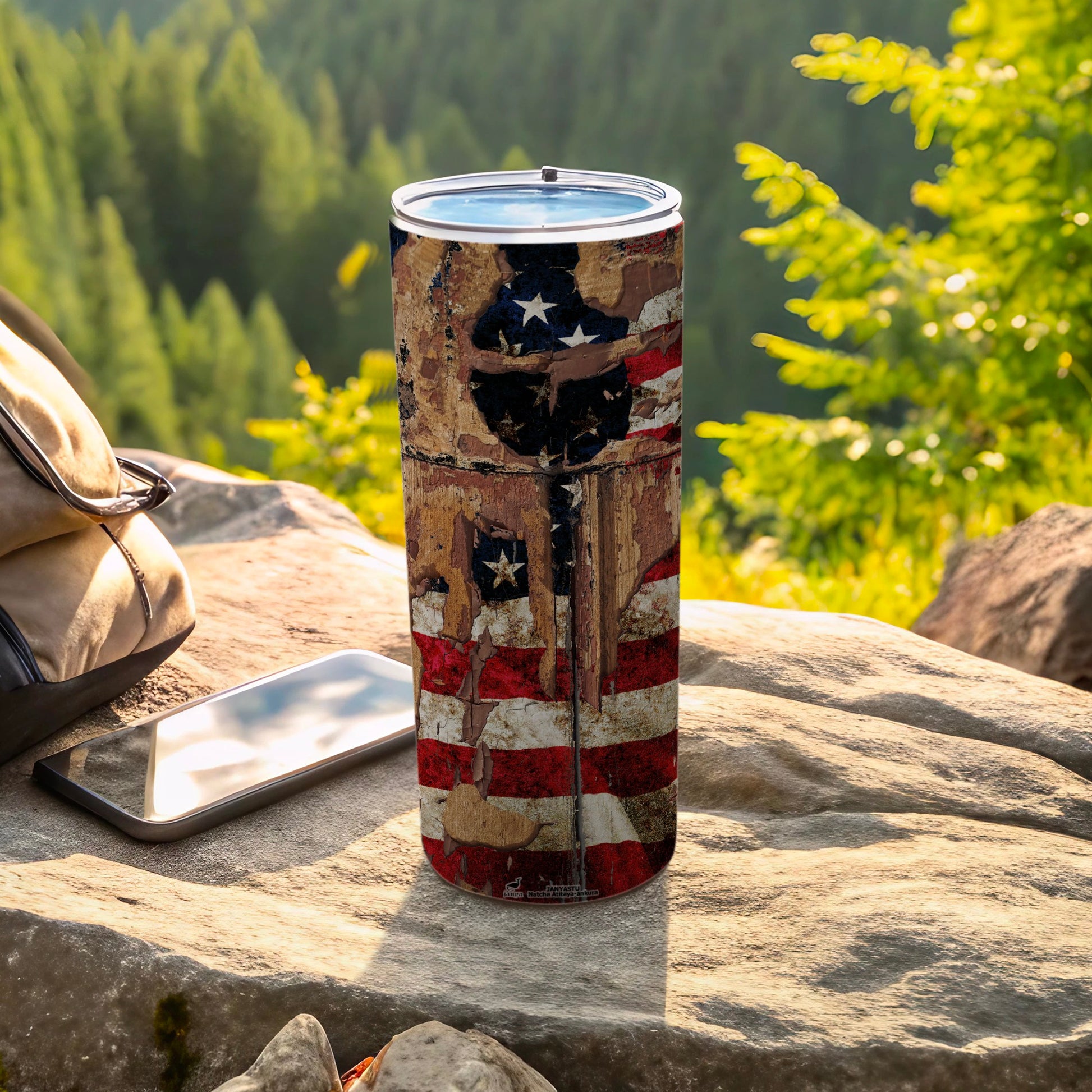 Distressed American flag tumbler with backpack and phone on rocks.