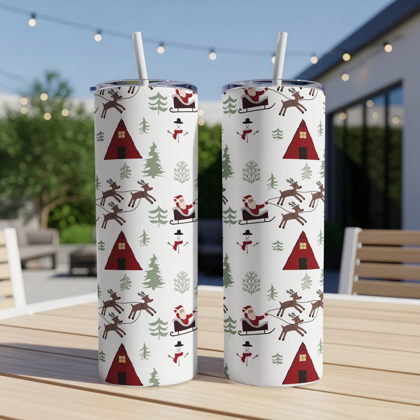 Two Christmas-themed tumblers with Santa, reindeer, snowmen, and a barn on a wooden table outdoors.