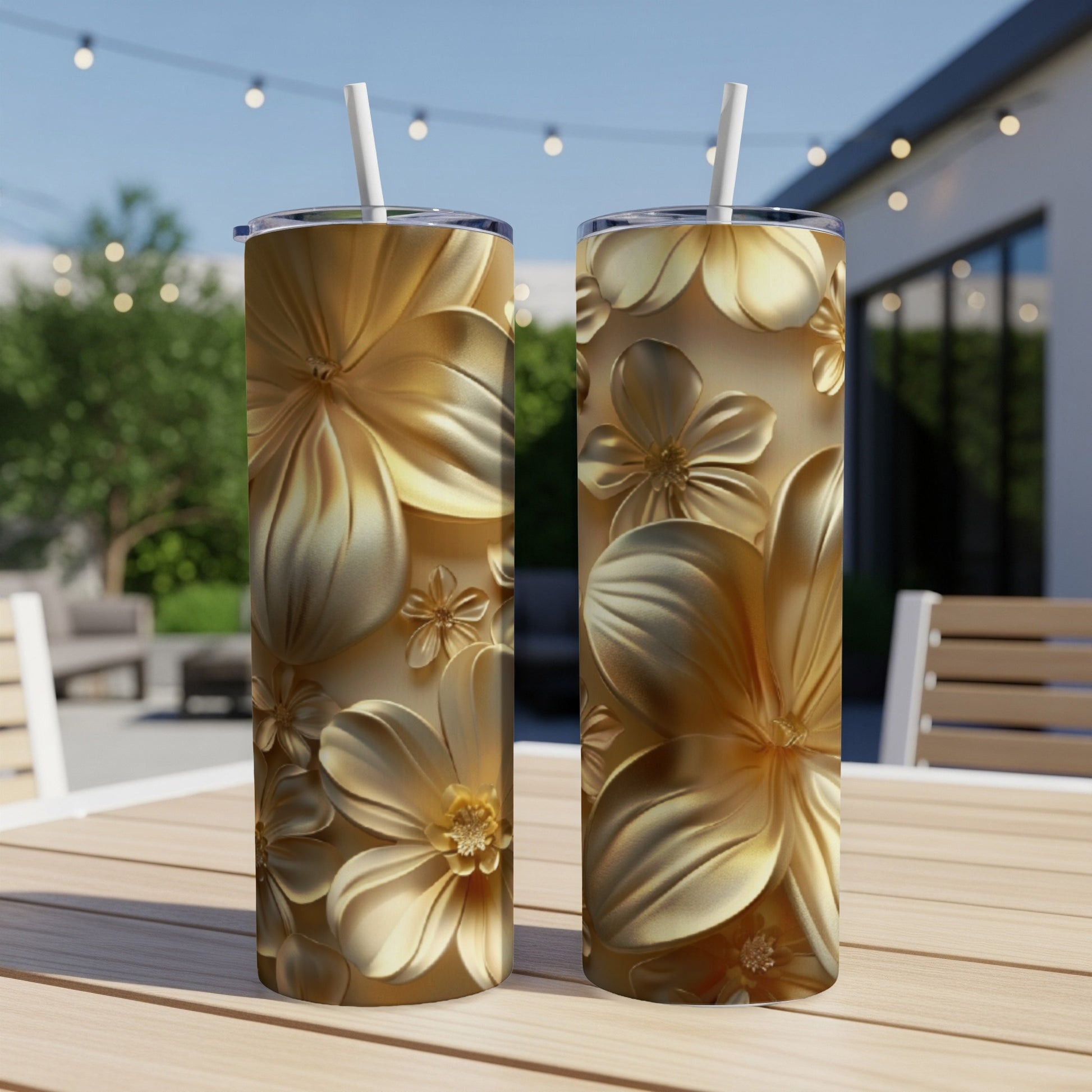 Two gold floral patterned tumblers with straws on a wooden table outdoors.