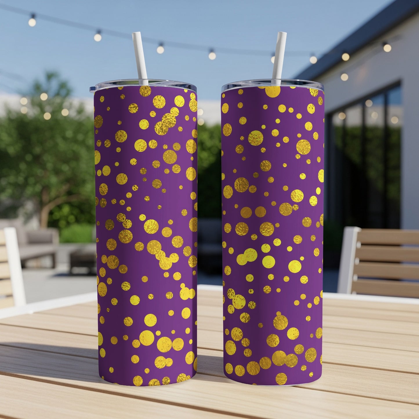 Two purple tumblers with gold polka dots and white straws on a wooden table outdoors.