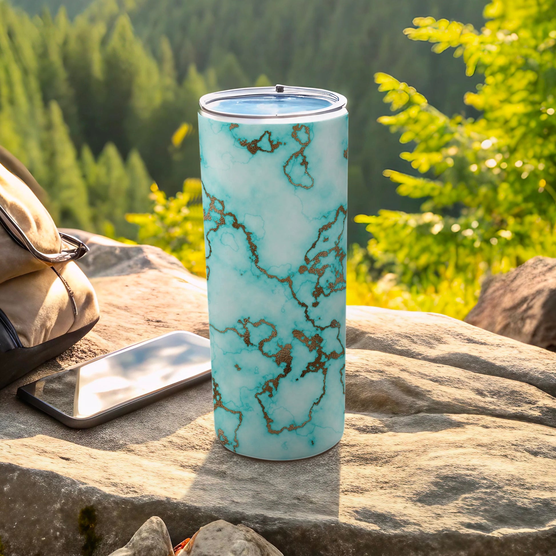 Teal marble tumbler with gold veins on a rock, with backpack and phone.
