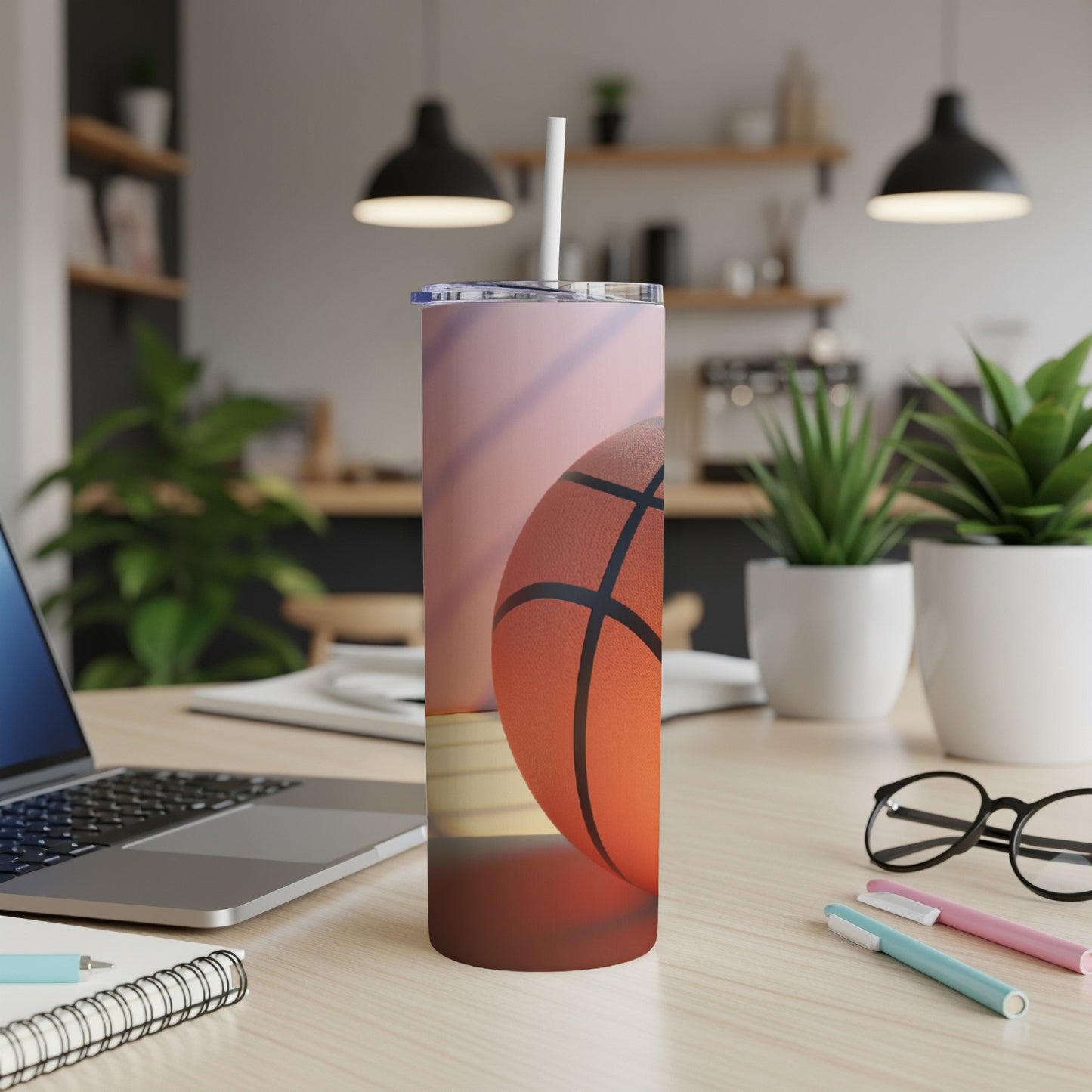 Basketball tumbler with straw on desk beside laptop and pens.