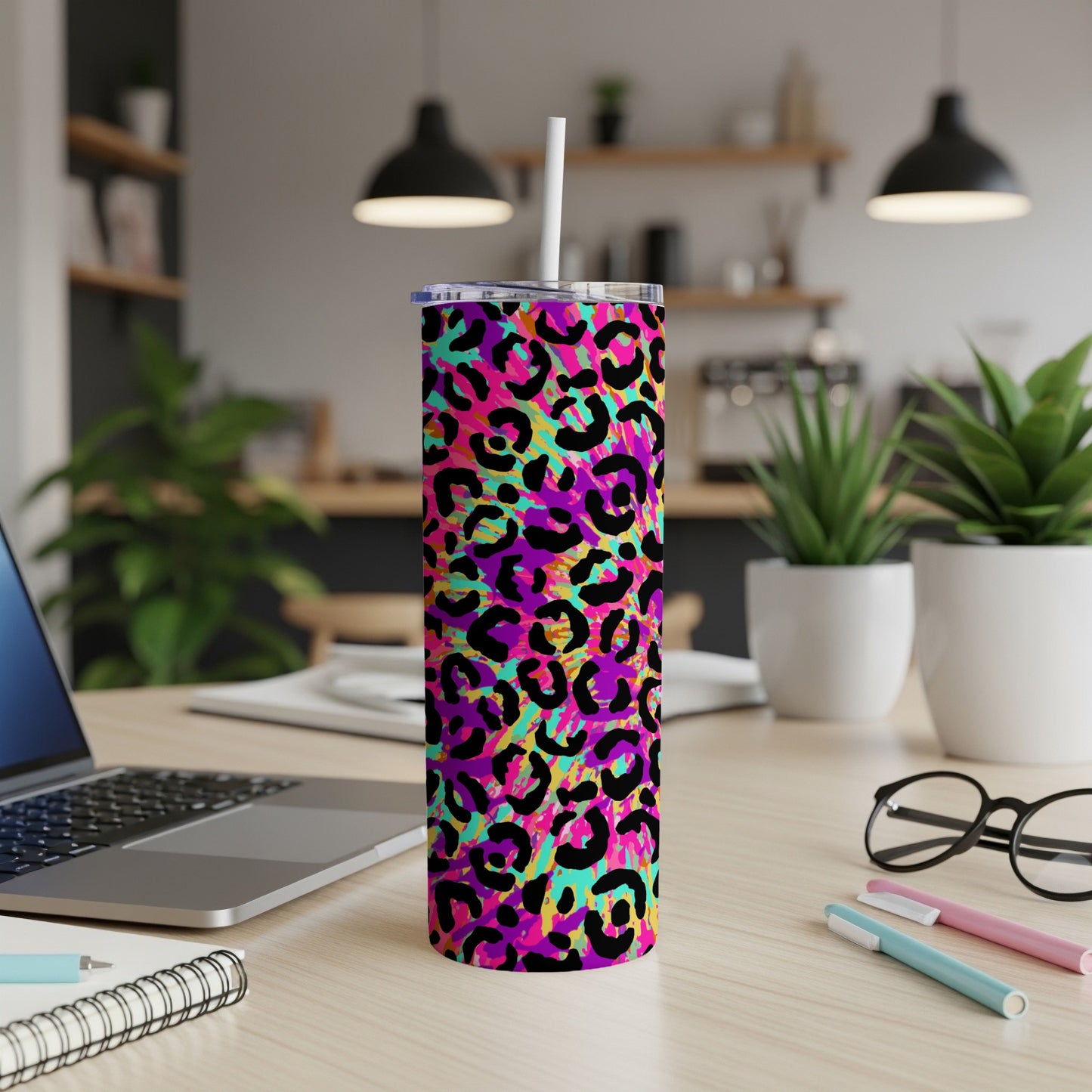 Tall tumbler with colorful leopard print and straw on a desk, laptop and glasses nearby.