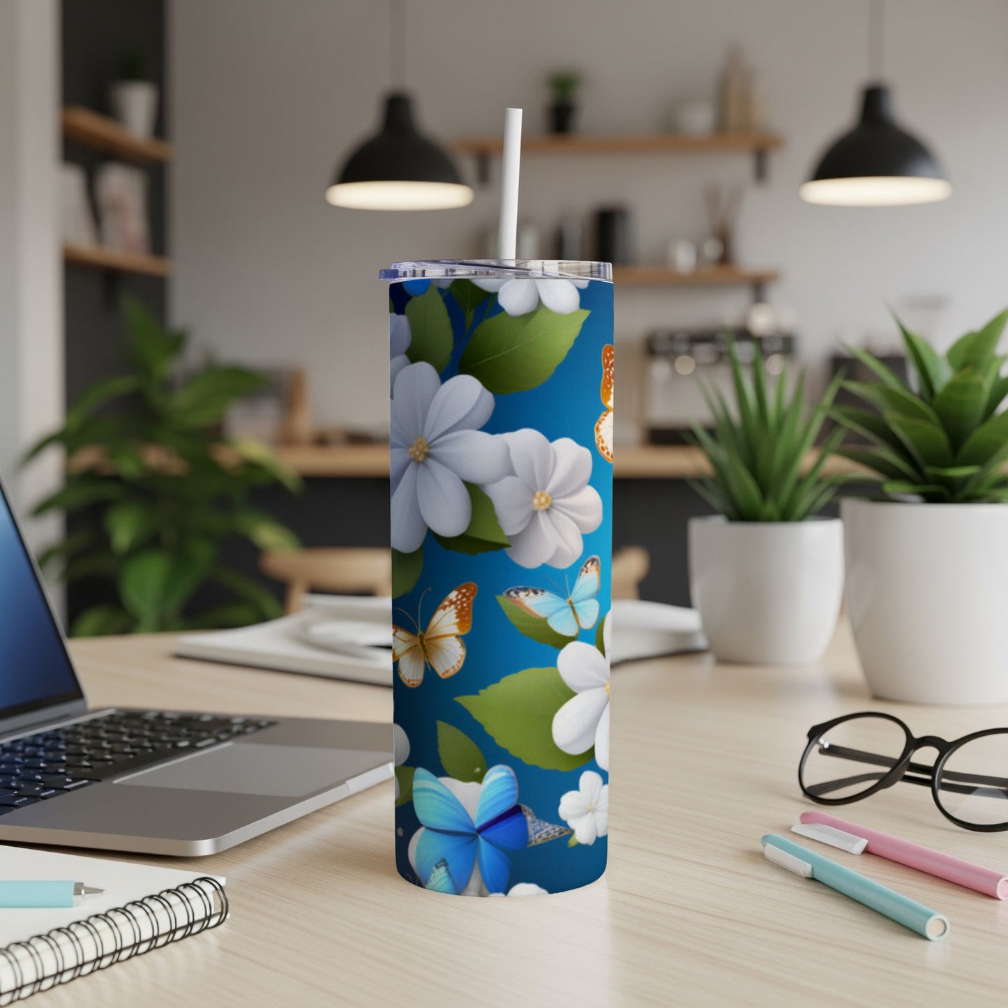 Blue tumbler with butterflies and white flowers, on a desk with laptop and pens.