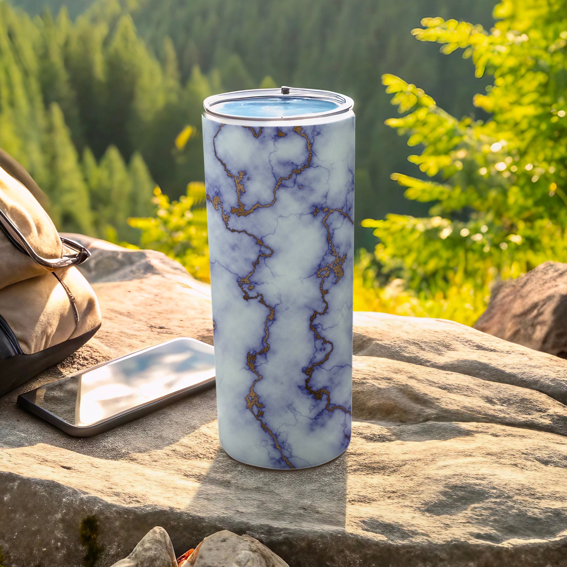 Marble-patterned tumbler with gold veining sits on rocks near a backpack and phone.