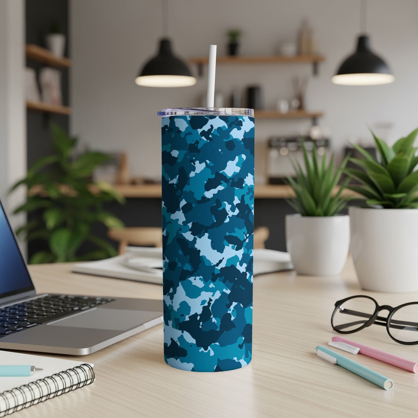 Blue camouflage tumbler with straw on a desk, next to laptop and plants.